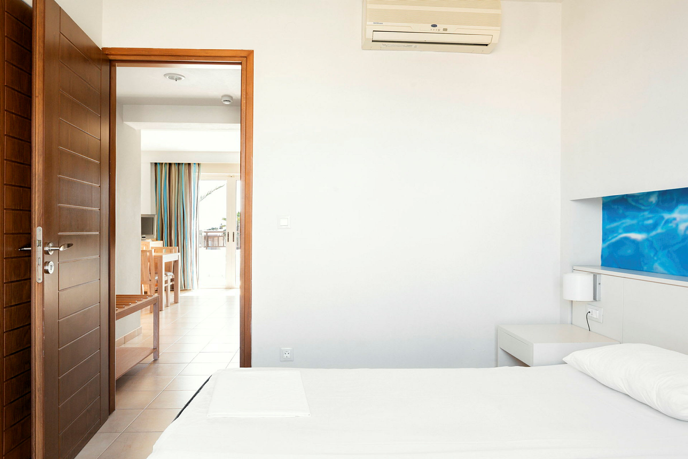Ett hotellrum i modern stil med vita väggar, en bäddad säng och dörr mot en ljus balkong. (AI-genererad text)