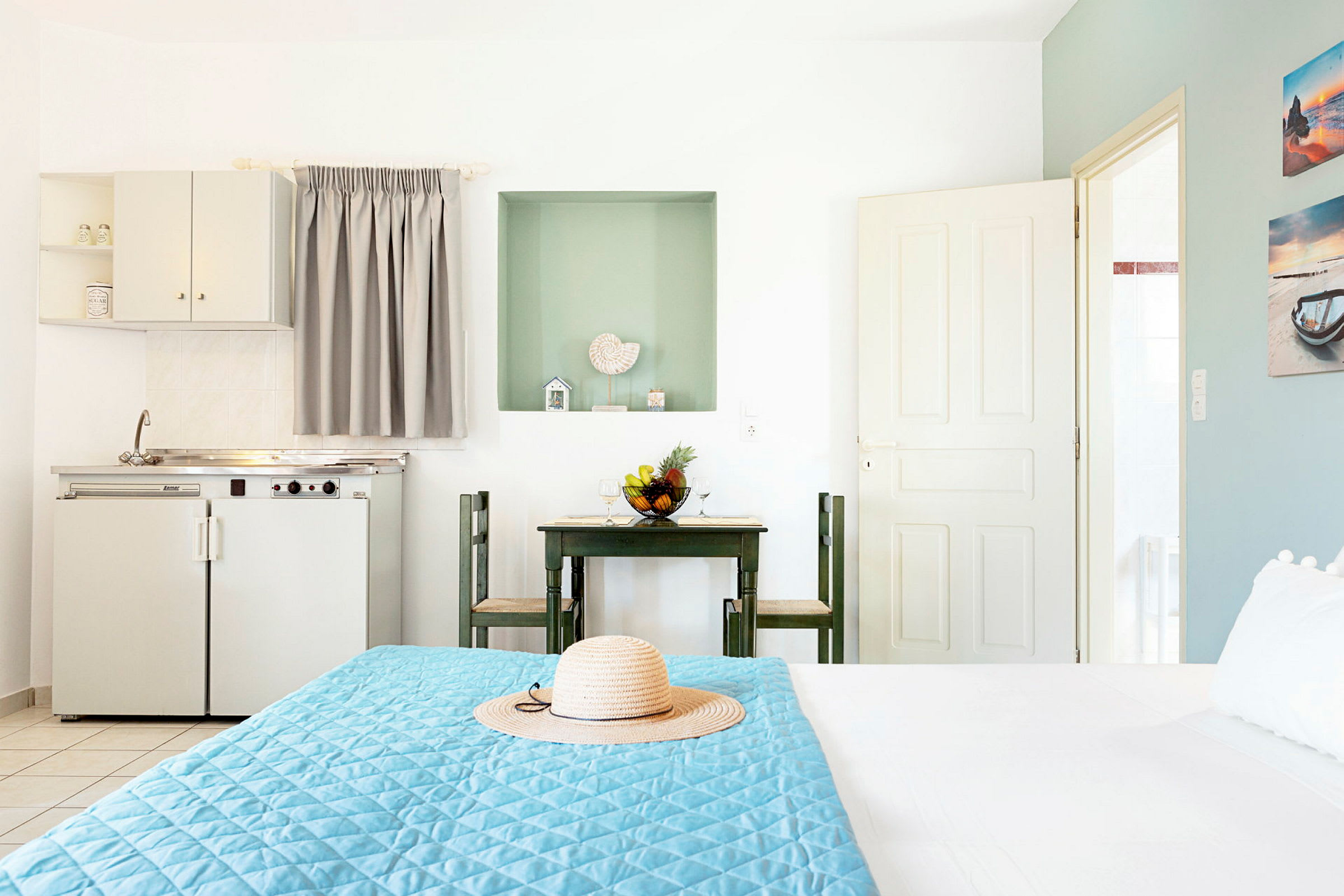 Ett hotellrum med en liten köksdel och matbord med två stolar, samt en säng med blå överkast och en stråhatt. (AI-genererad text)