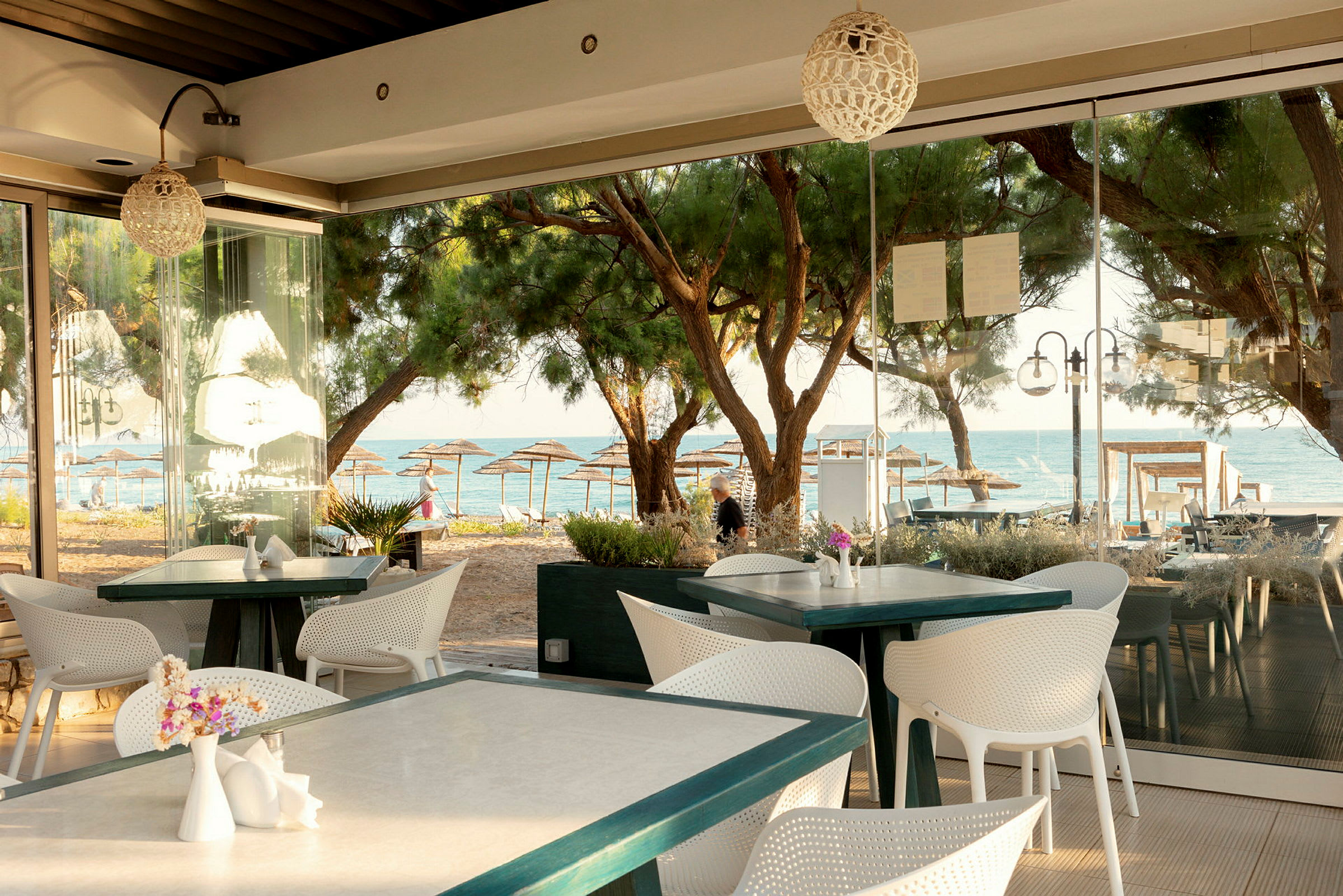 Ett café vid stranden med vita möbler och bord med utsikt över träd, stranden och havet. (AI-genererad text)