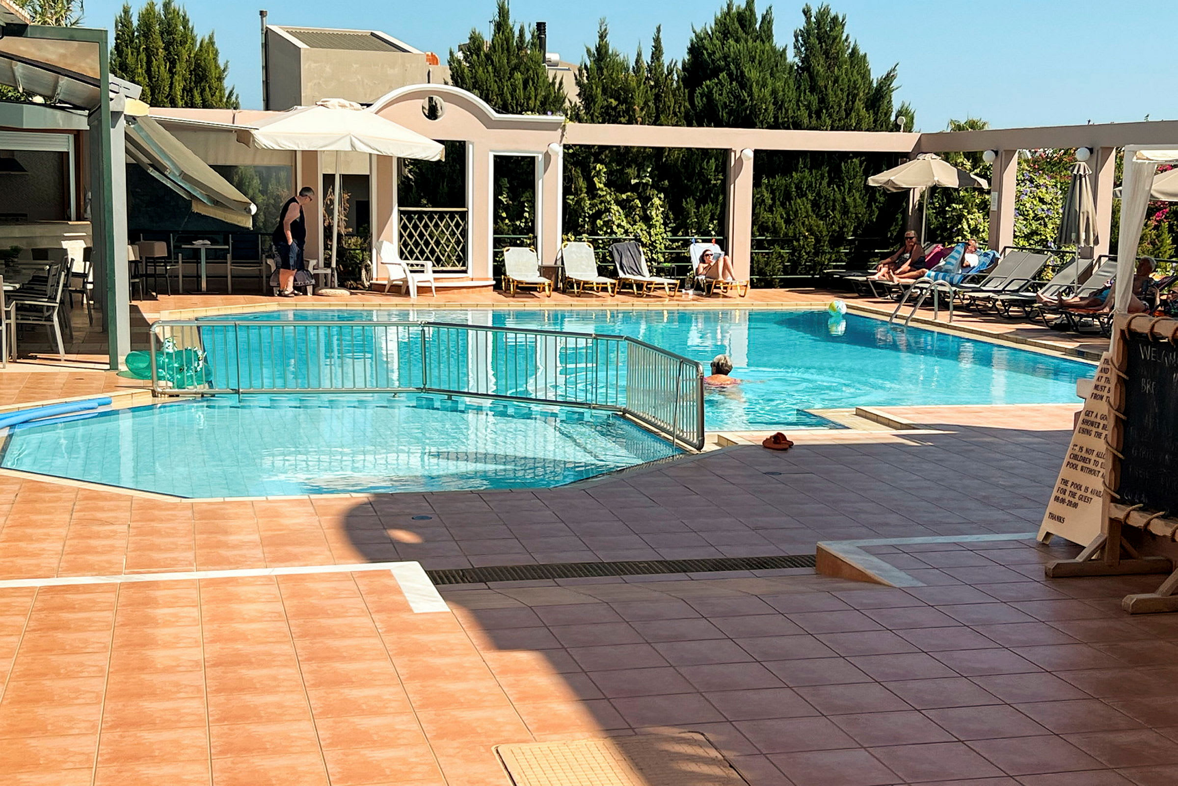 Utomhuspool med solstolar och parasoll vid sidan av, samt människor som badar och solar. (AI-genererad text)