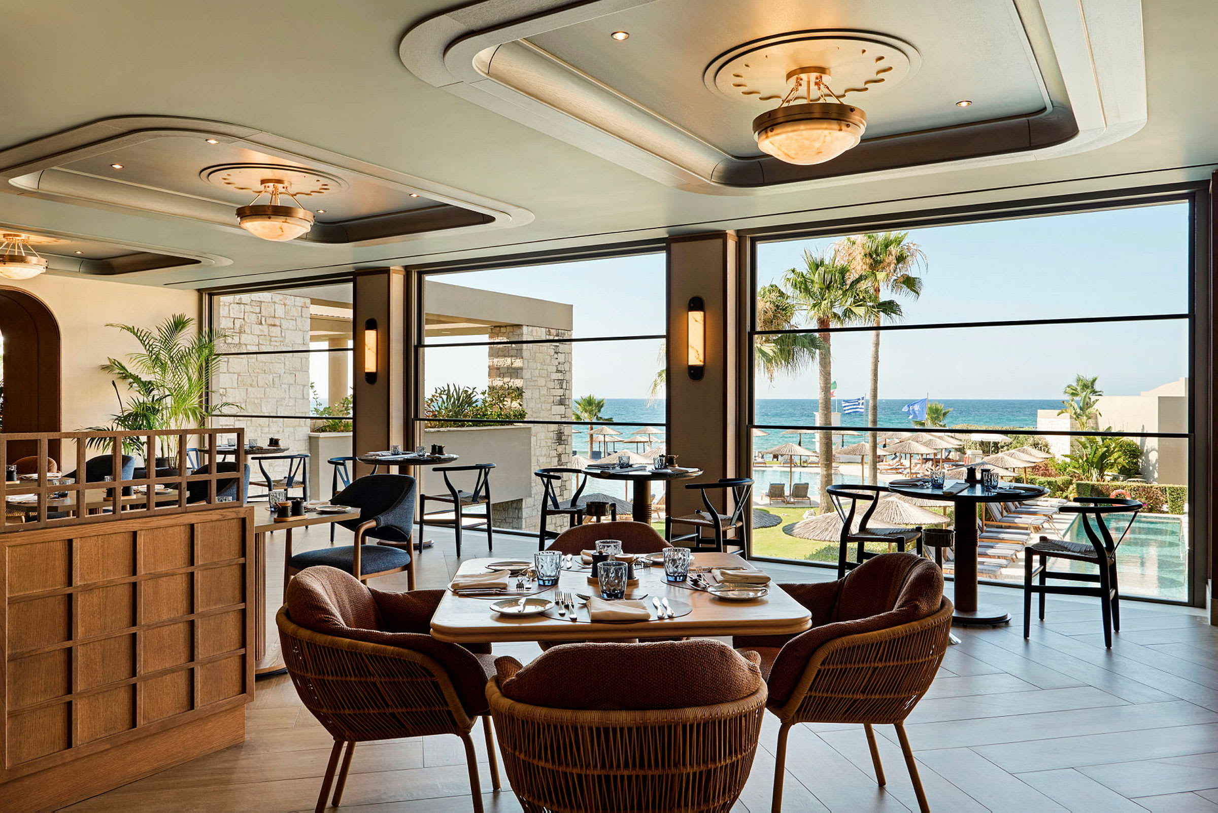 Interiör av en restaurang med stora fönster som vetter mot en havsutsikt, palmträd och poolområdet utanför. (AI-genererad text)