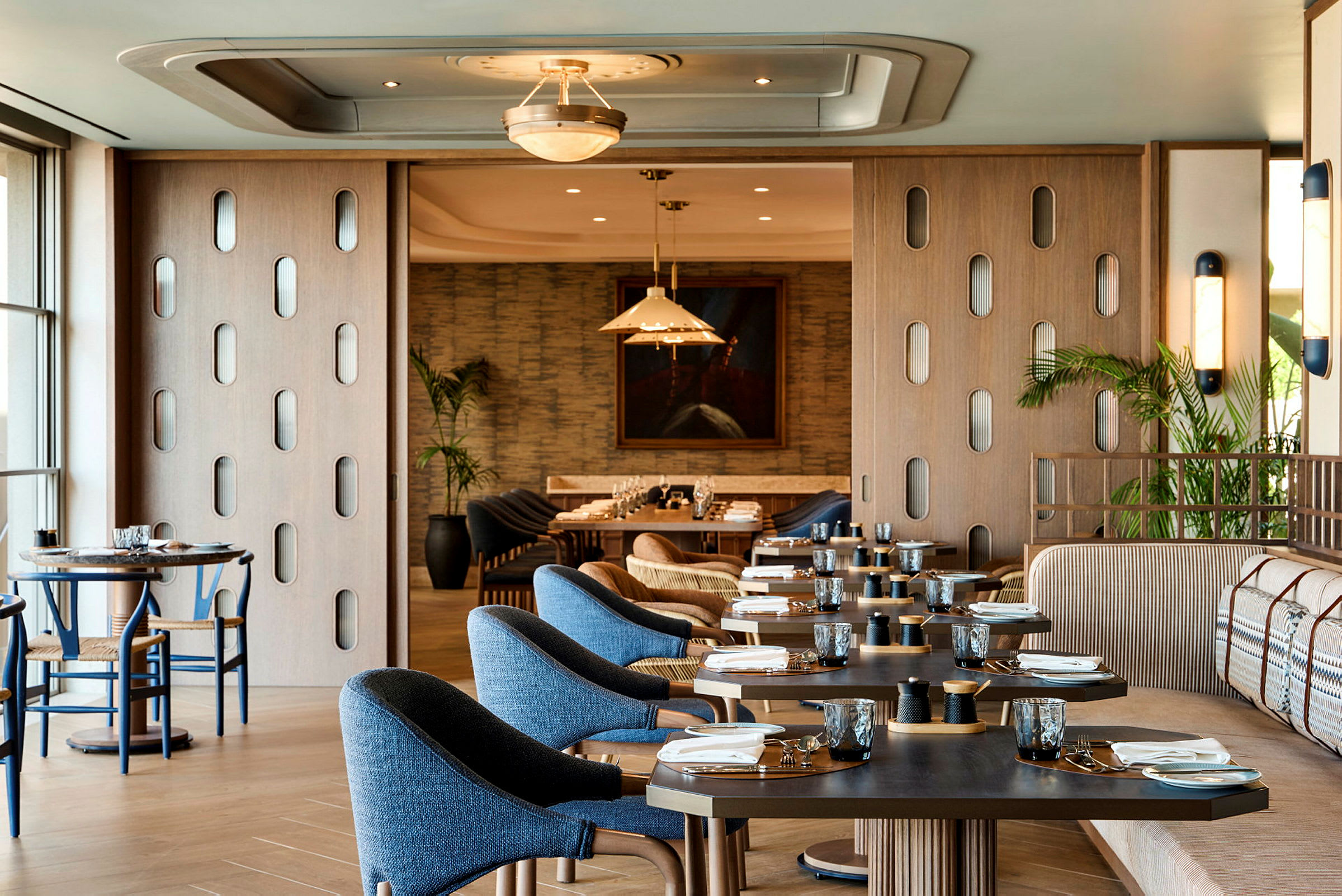 Inredningen i en modern restaurang med träbord och stolar, blå klädsel och öppet mot ett annat rum med en sittgrupp. (AI-genererad text)