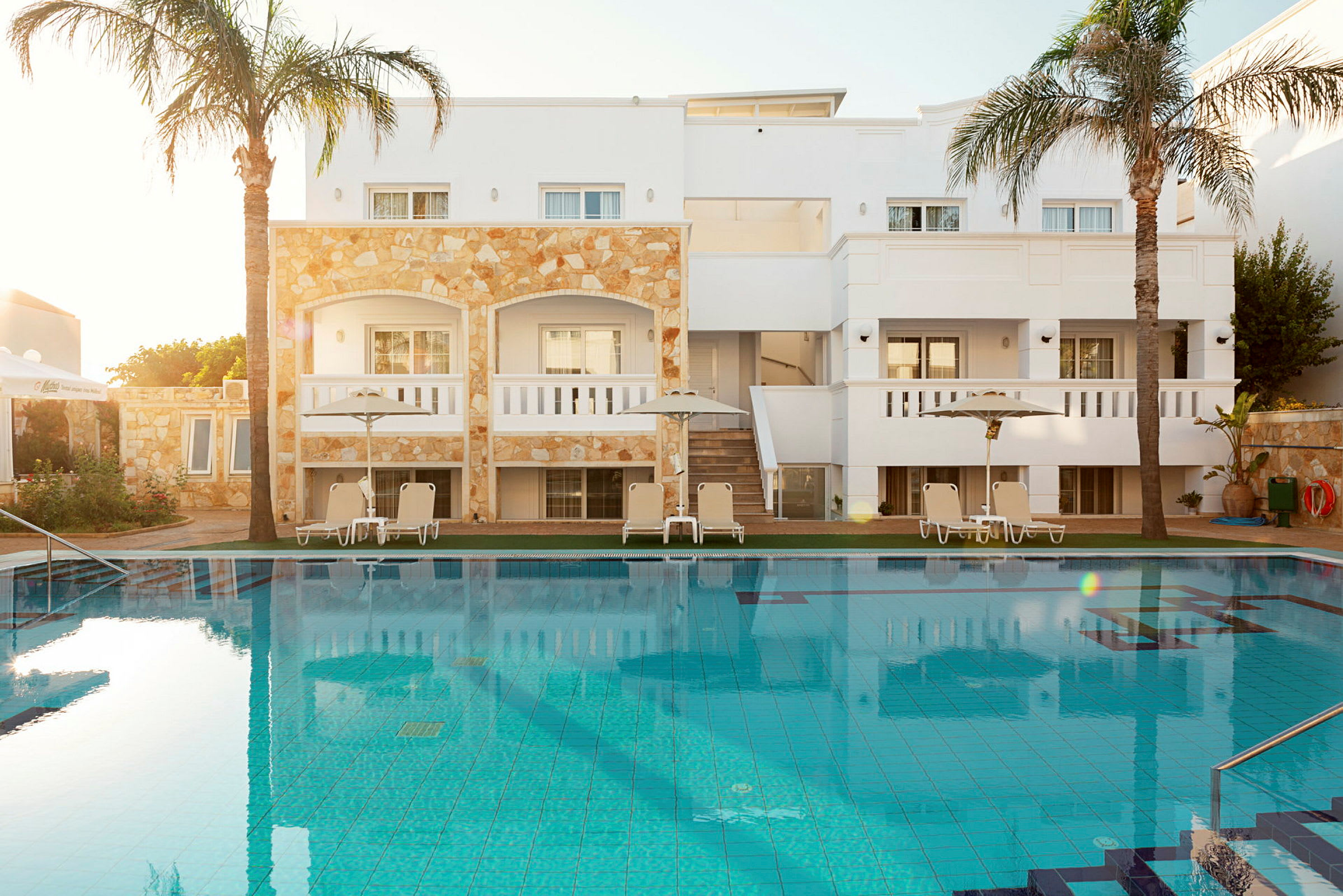 Bild av ett hotellområde med en swimmingpool i förgrunden och solstolar under parasoller vid poolkanten framför en byggnad. (AI-genererad text)