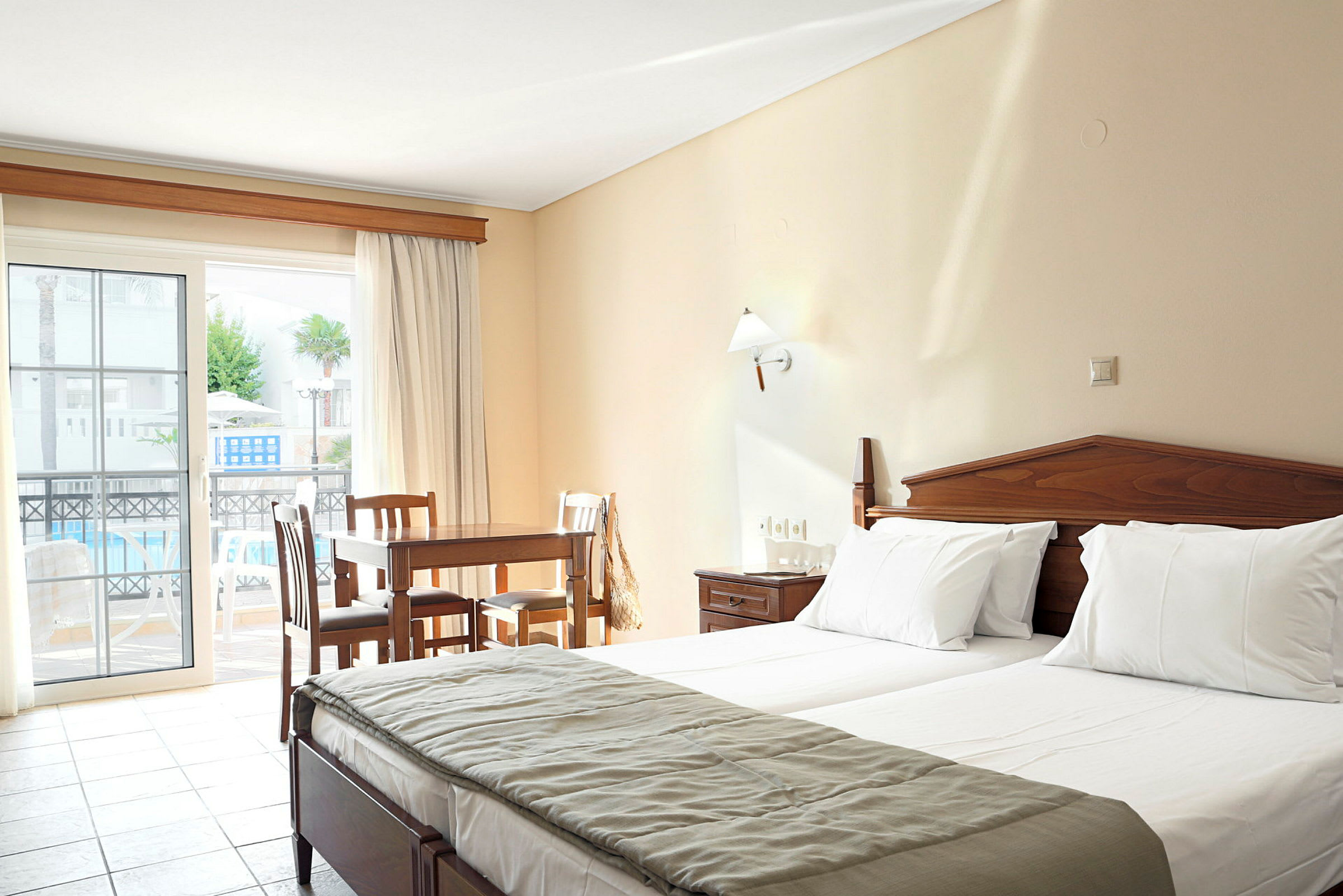 Ett hotellrum med två sängar, ett träbord och stolar, samt en balkong med utsikt över poolområdet. (AI-genererad text)