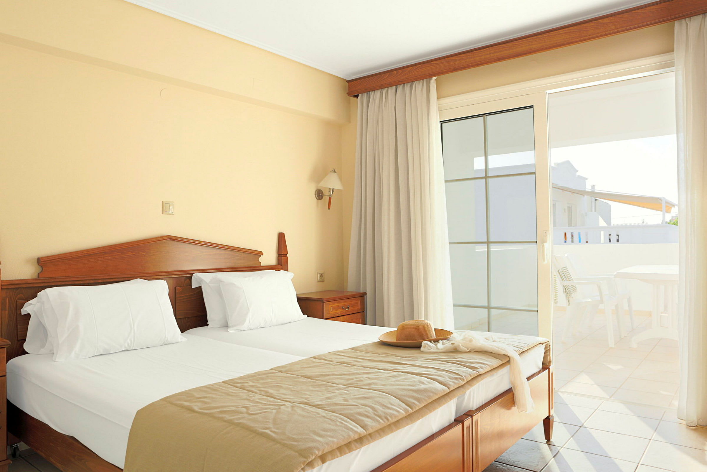 Ett hotellrum med en dubbelsäng, ljusa gardiner och en balkong med utemöbler. (AI-genererad text)
