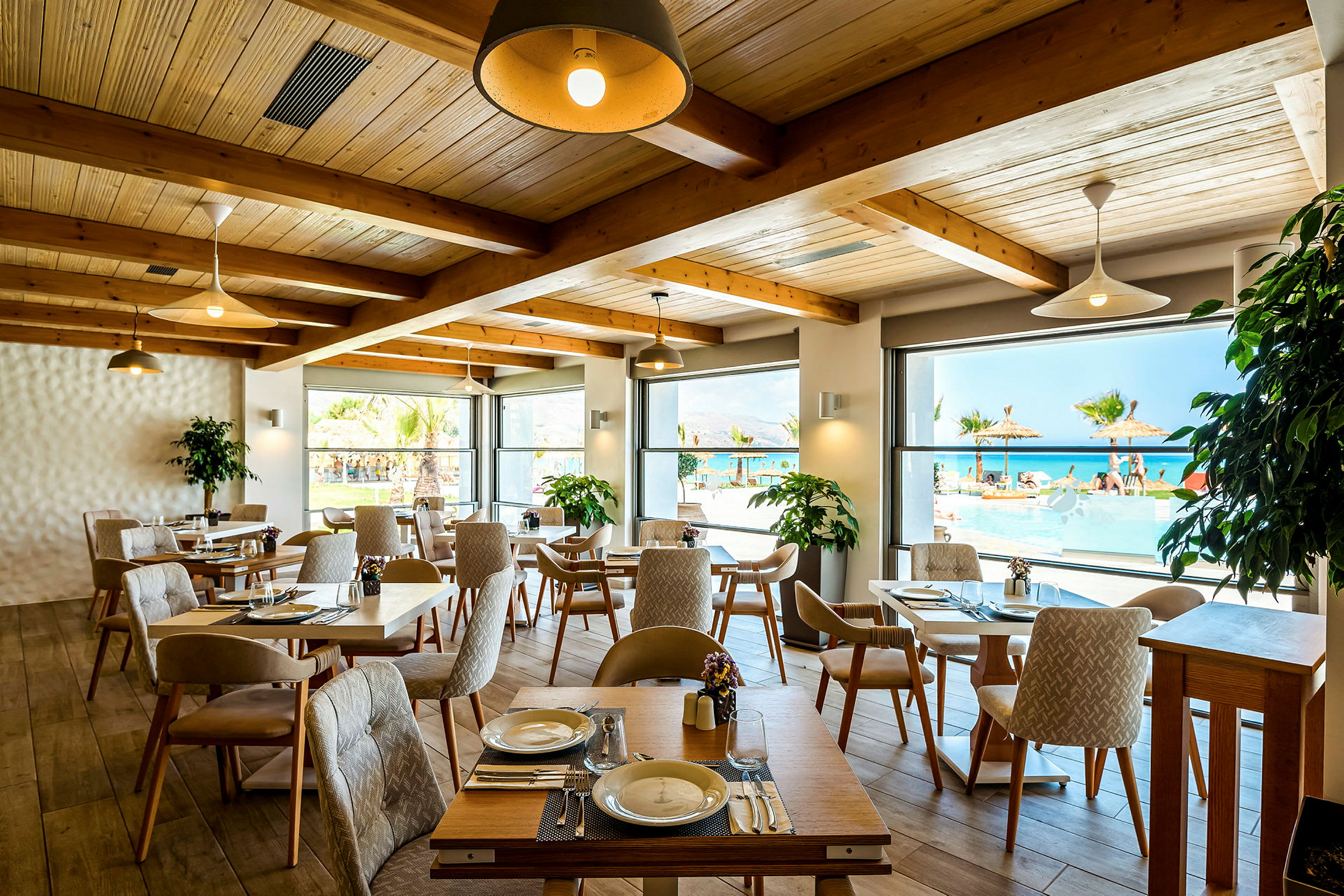 En restaurang med bord och stolar inomhus och utsikt över pool och hav genom fönstren. (AI-genererad text)
