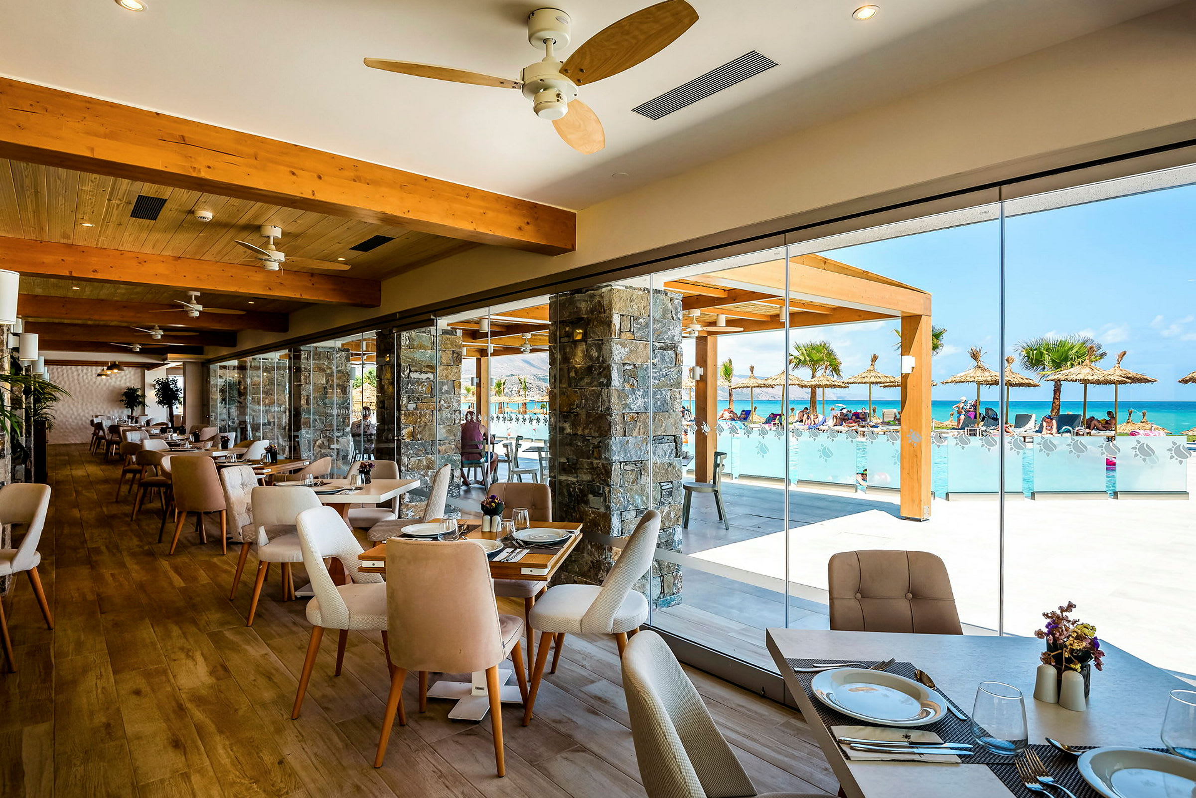 Restaurang med glasväggar som vetter mot en pool och palmbeklädda parasoller. (AI-genererad text)