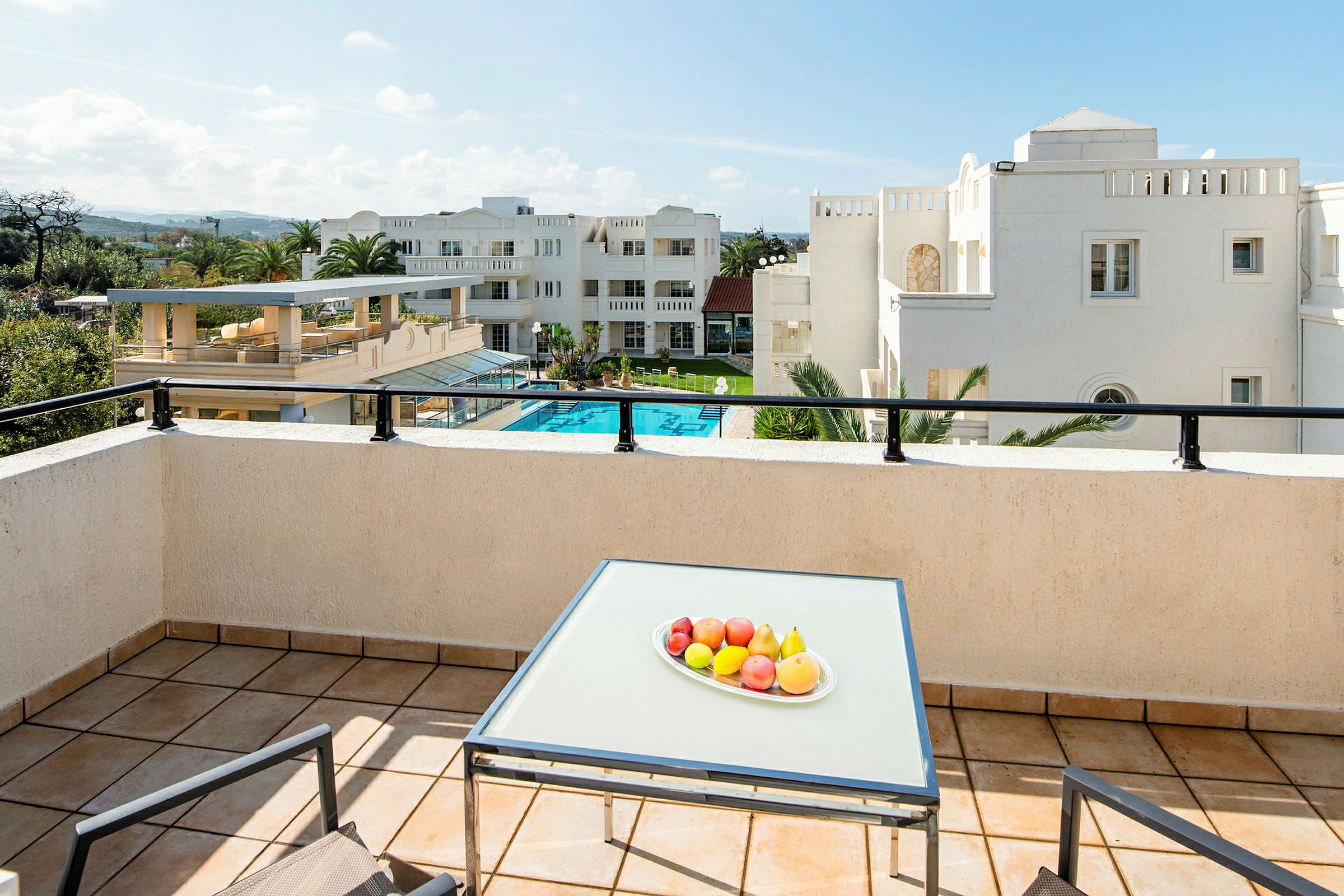 En balkong med ett bord på vilket en skål med frukt står. I bakgrunden syns en swimmingpool och vita byggnader. (AI-genererad text)
