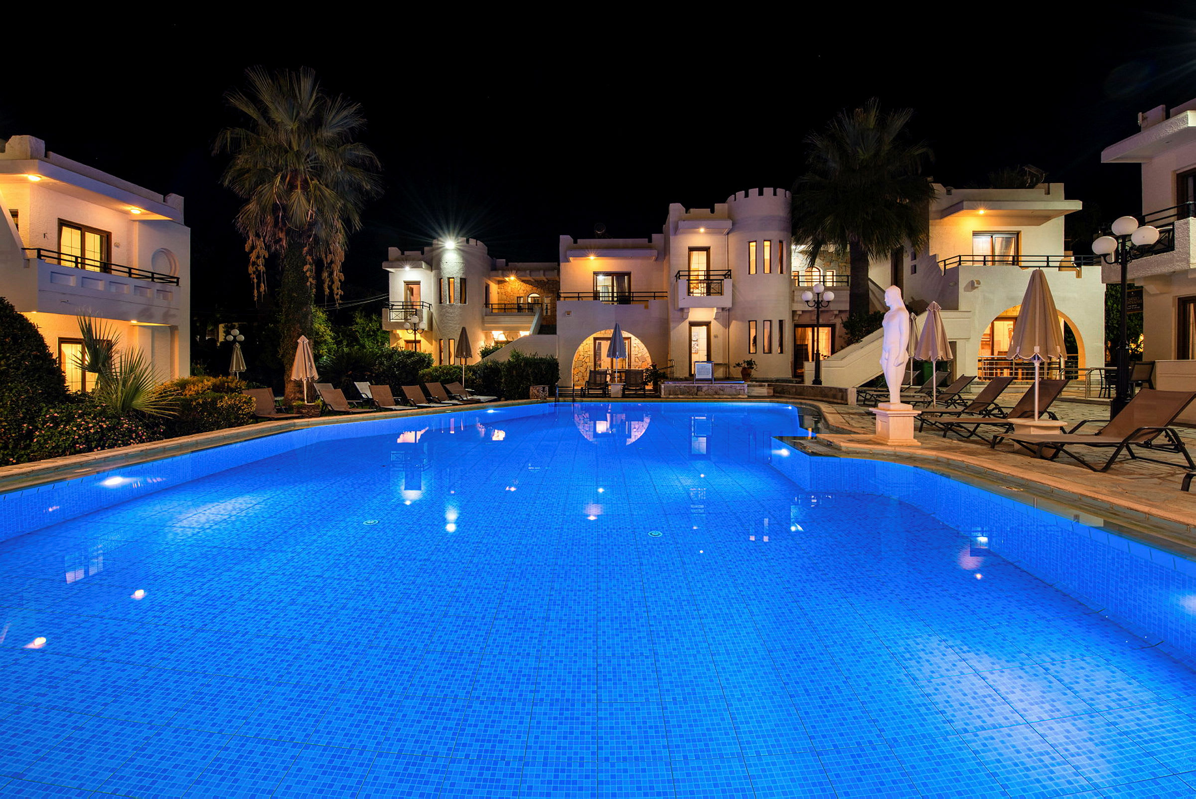 En upplyst pool framför hotellbyggnader i medeltida stil under natten. (AI-genererad text)