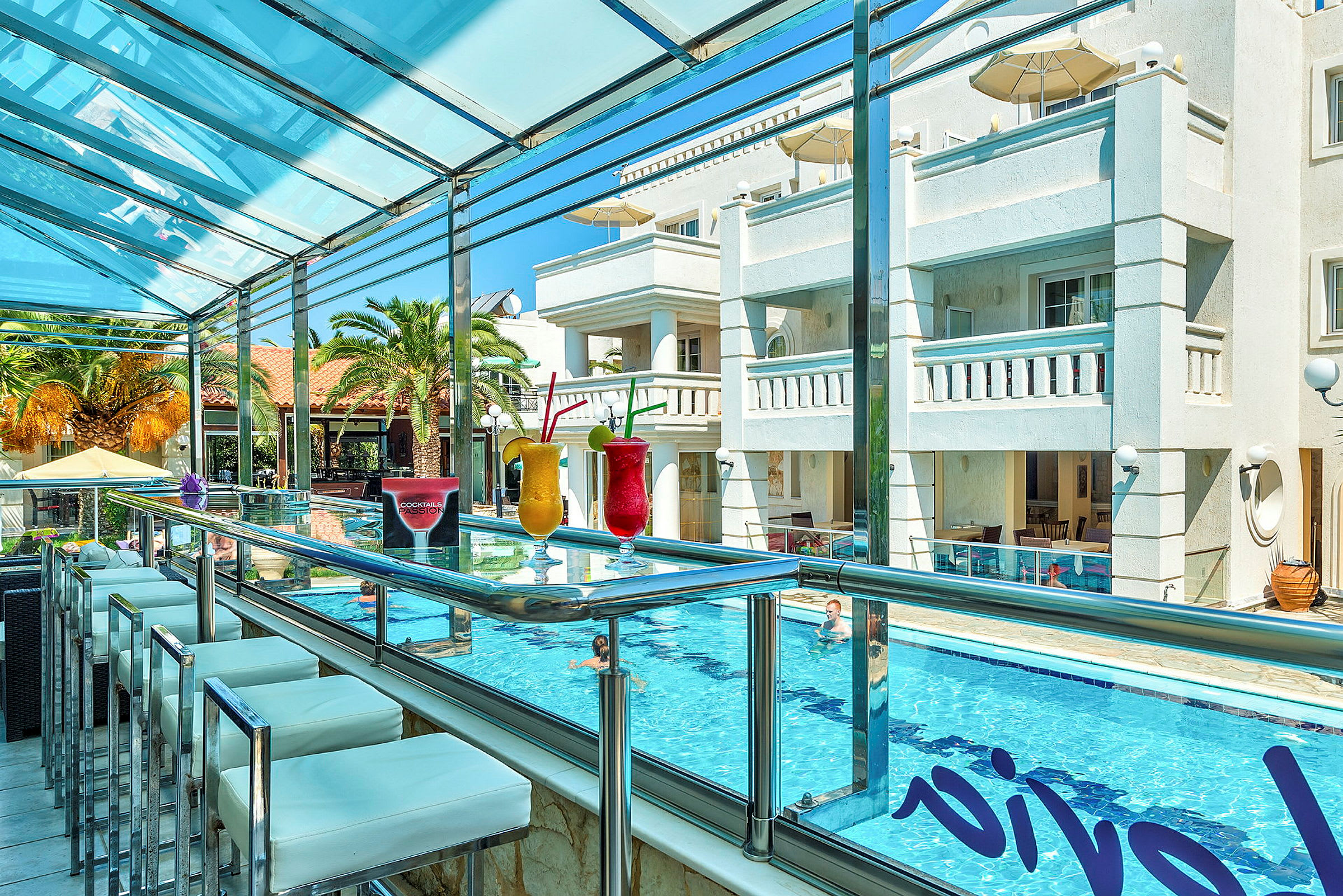 Bild av en utomhusbar med glasräcke, där färgglada cocktails är upplagda på disken. I bakgrunden syns en pool och en hotellbyggnad. (AI-genererad text)