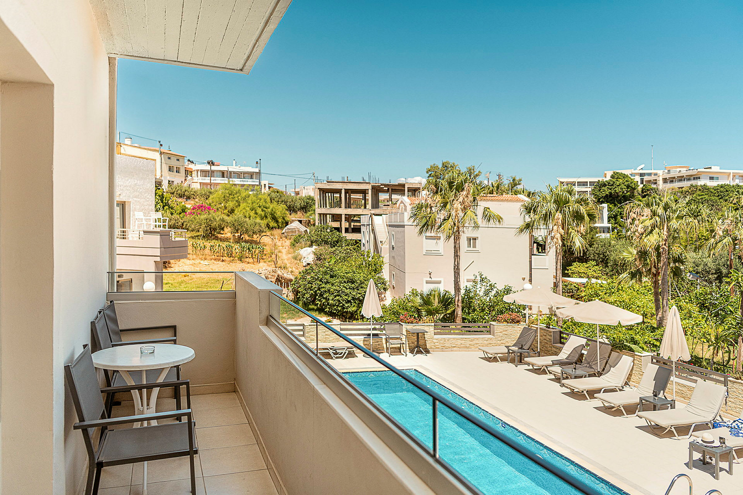 Bild av en balkong med utsikt över en pool och omgivande byggnader och grönska. Solstolar med parasoller står uppställda bredvid poolen. (AI-genererad text)