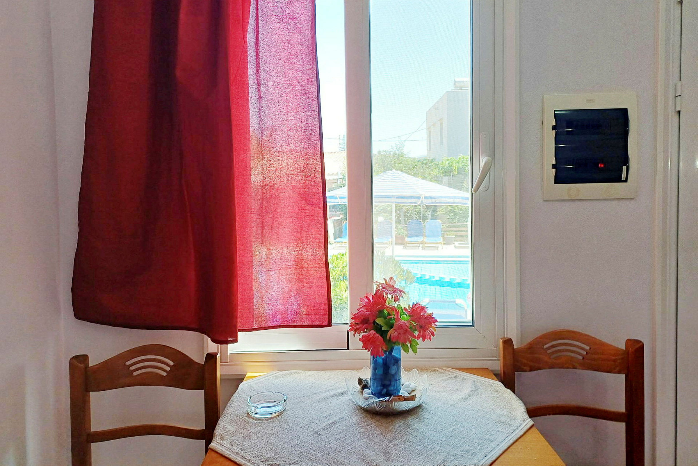 En interiör med ett bord med en vas med blommor framför ett fönster med utsikt mot en pool. (AI-genererad text)