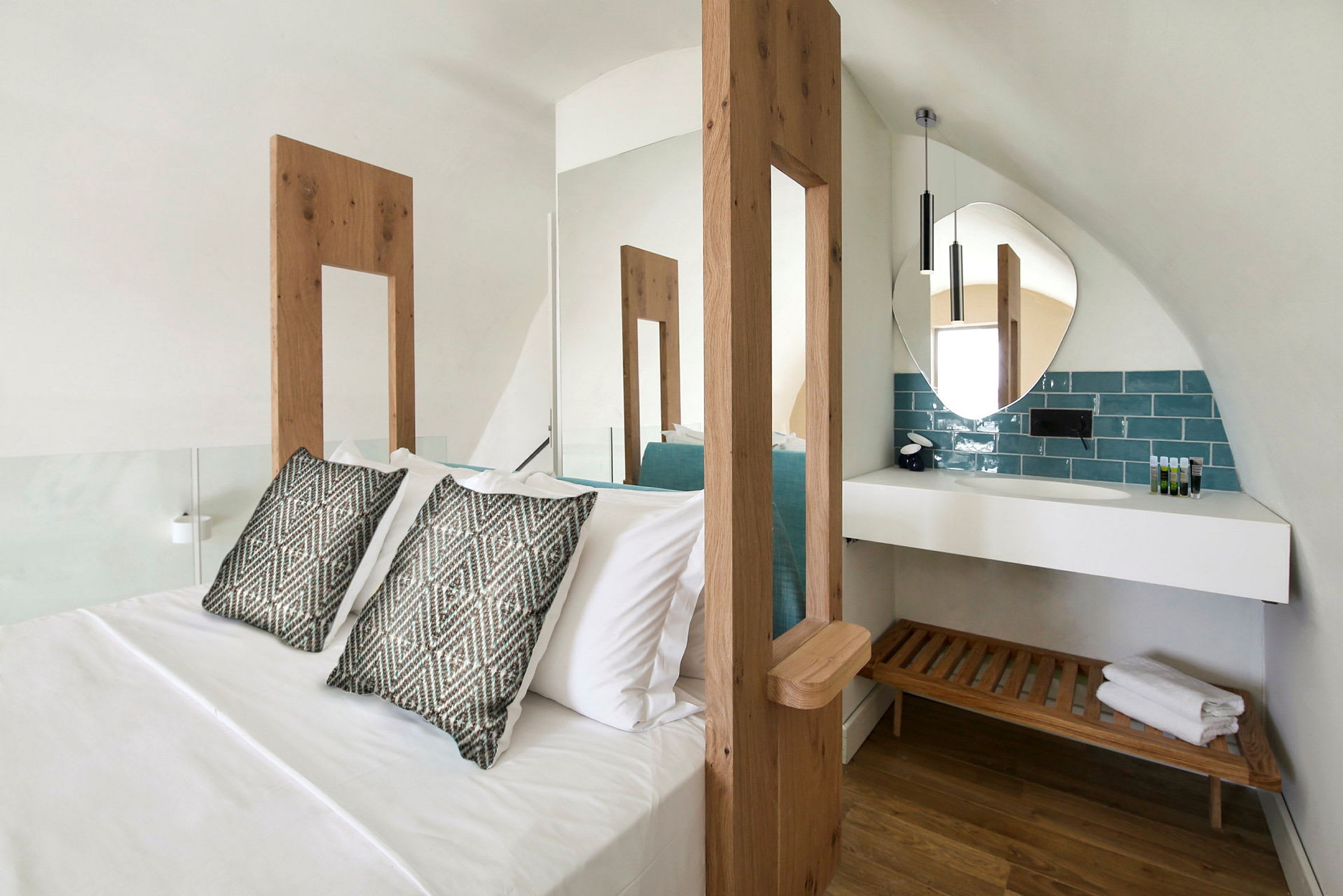 Ett hotellrum med en stor säng, två prydnadskuddar, och ett modernt badrumsområde med en rund spegel och en träbänk under en vit hylla. (AI-genererad text)