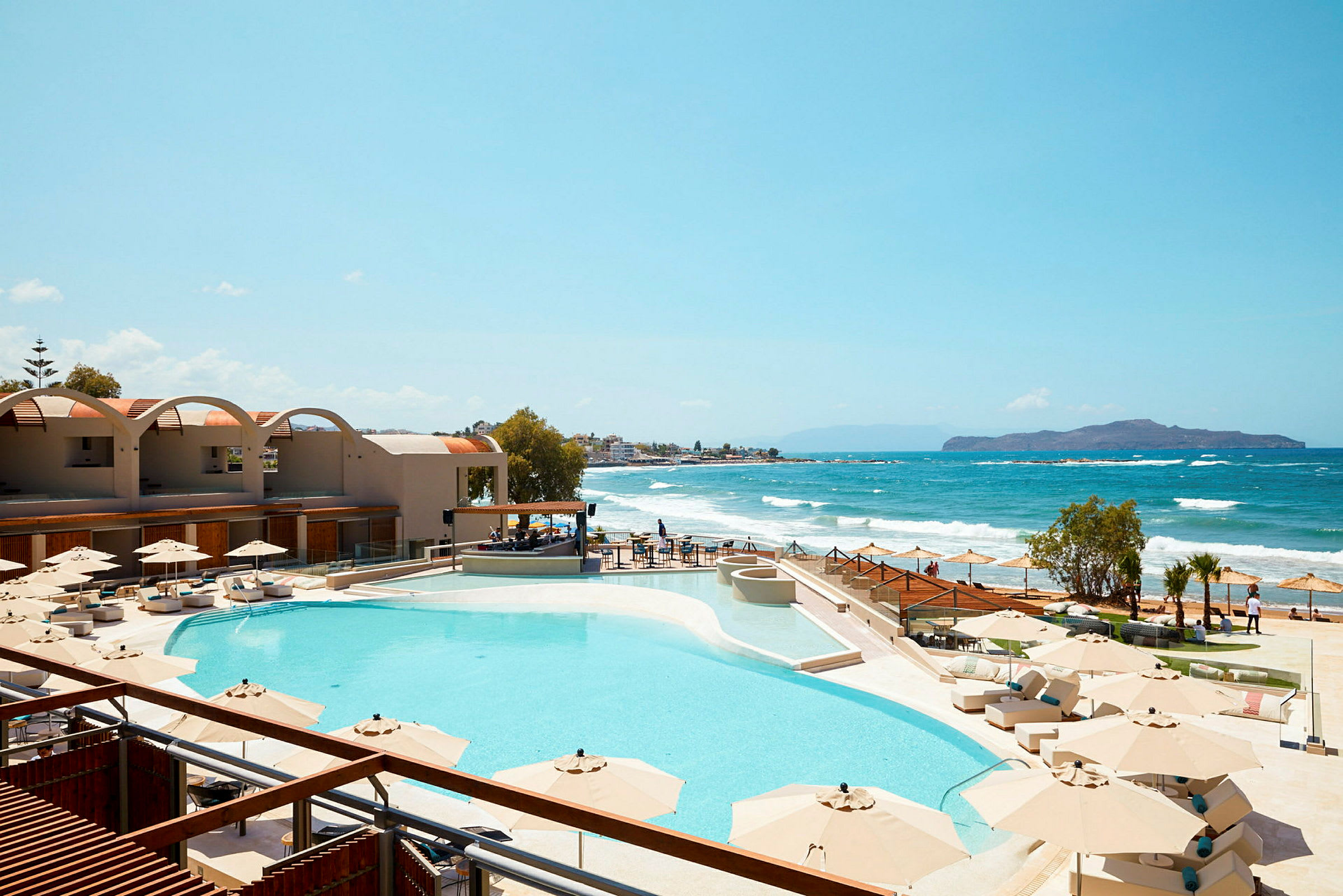 En pool med solparasoller vid en strand med vågor och utsikt över havet. (AI-genererad text)