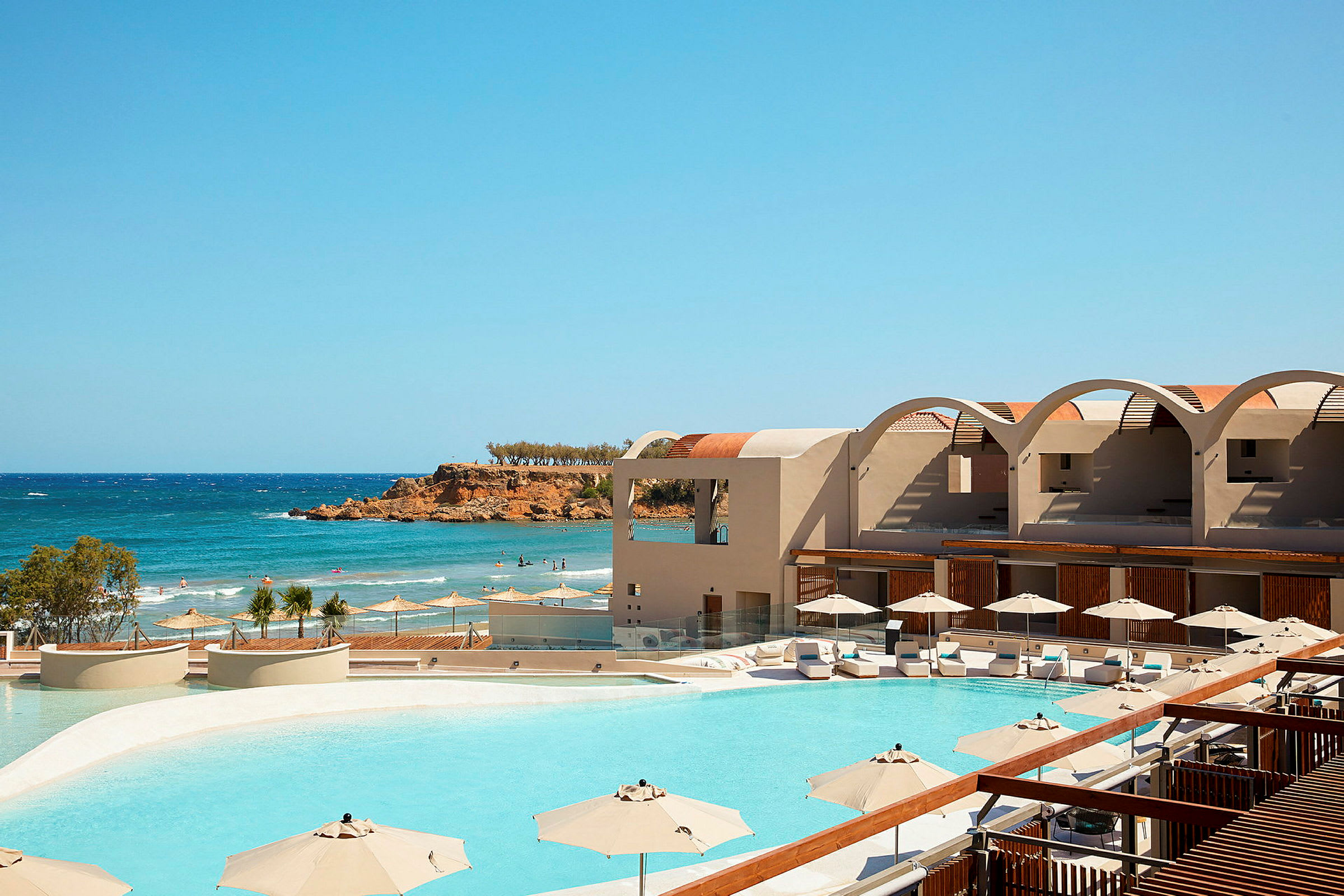 En hotellpool med solsängar och parasoller, med havet och en klippa i bakgrunden. (AI-genererad text)