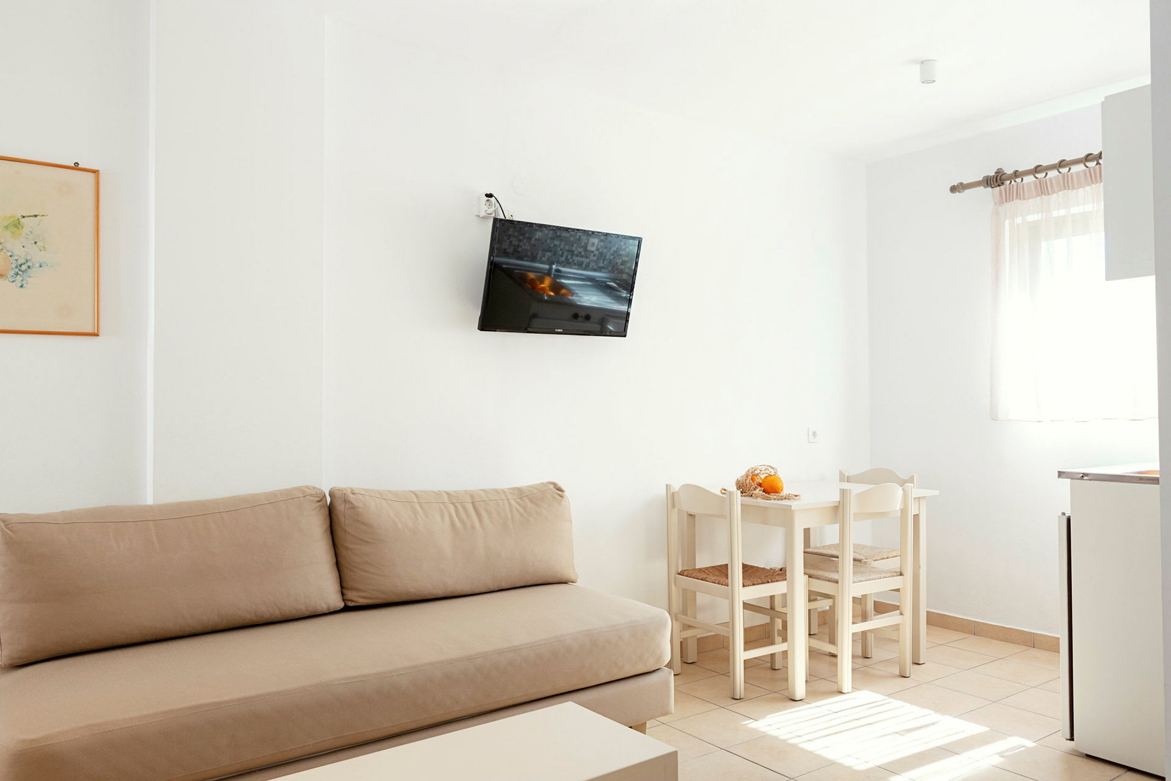 Ett ljust rum med beige soffa, vägghängd TV, vit matgrupp med stolar och ett fönster med ljusa gardiner. (AI-genererad text)