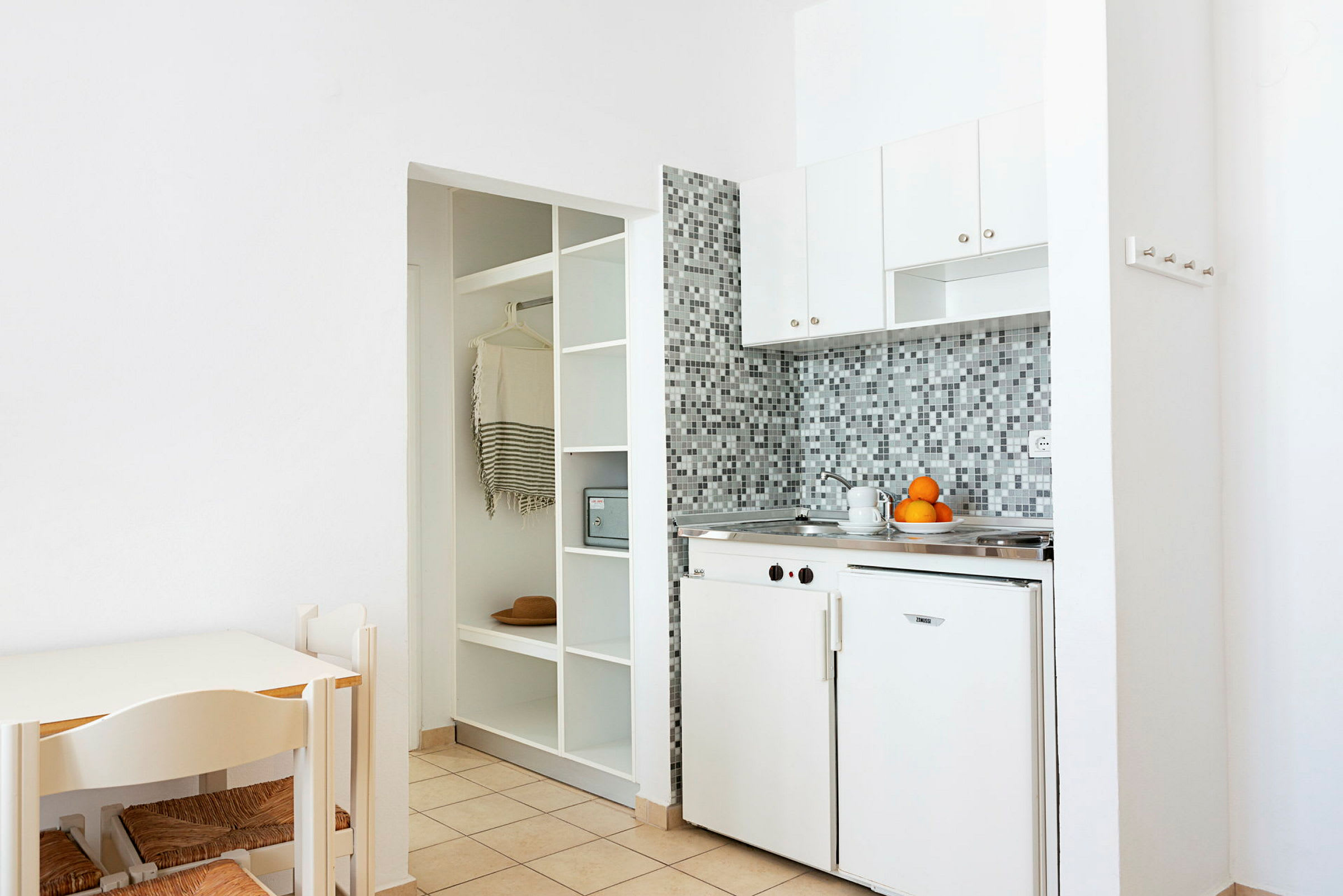 Ett kök med vita skåp, mosaik kakel och en minifrys; till vänster en matplats med stolar och ett bord. (AI-genererad text)