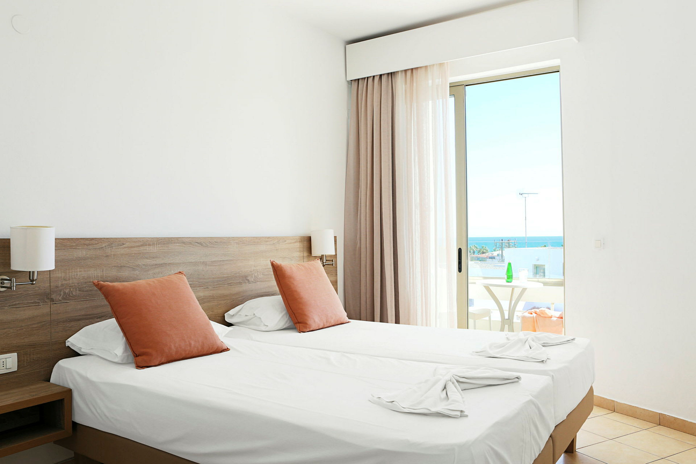 Hotellrum med två enkelsängar, orange kuddar och en balkong med havsutsikt. (AI-genererad text)