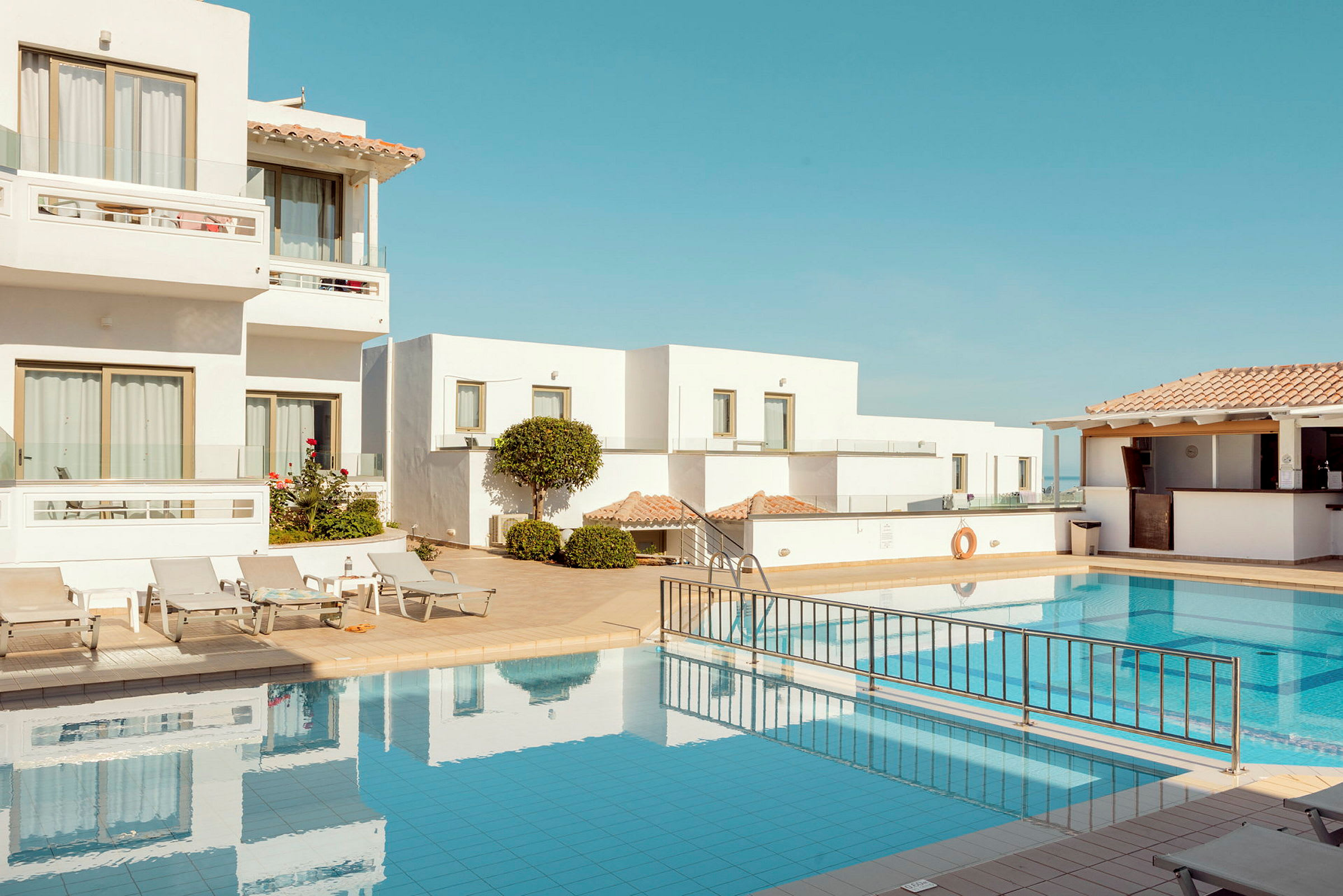 Vitmålade byggnader kring en pool med solstolar och klarblå himmel. (AI-genererad text)
