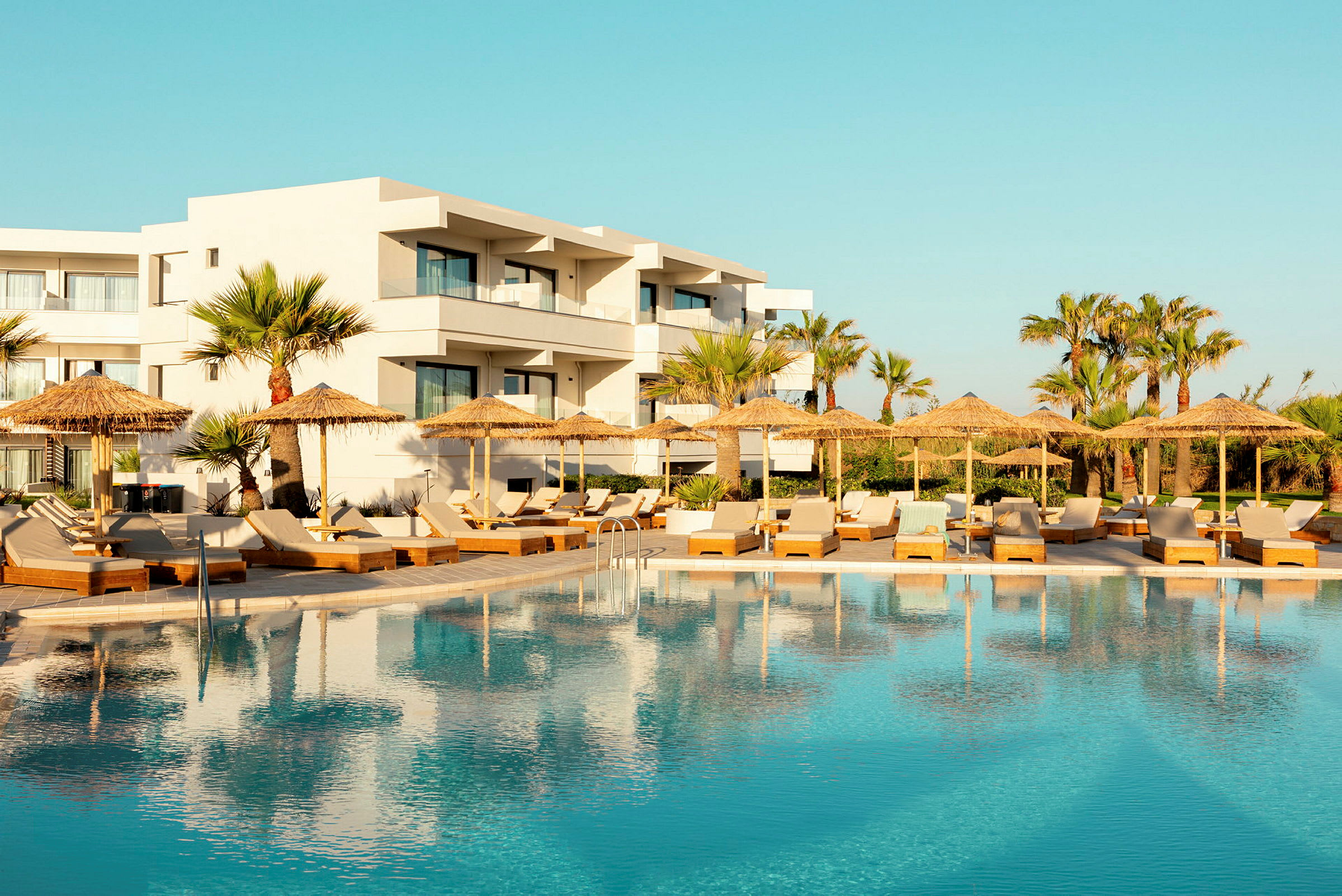 Hotell med vit fasad bakom en swimmingpool och solstolar med parasoller. (AI-genererad text)