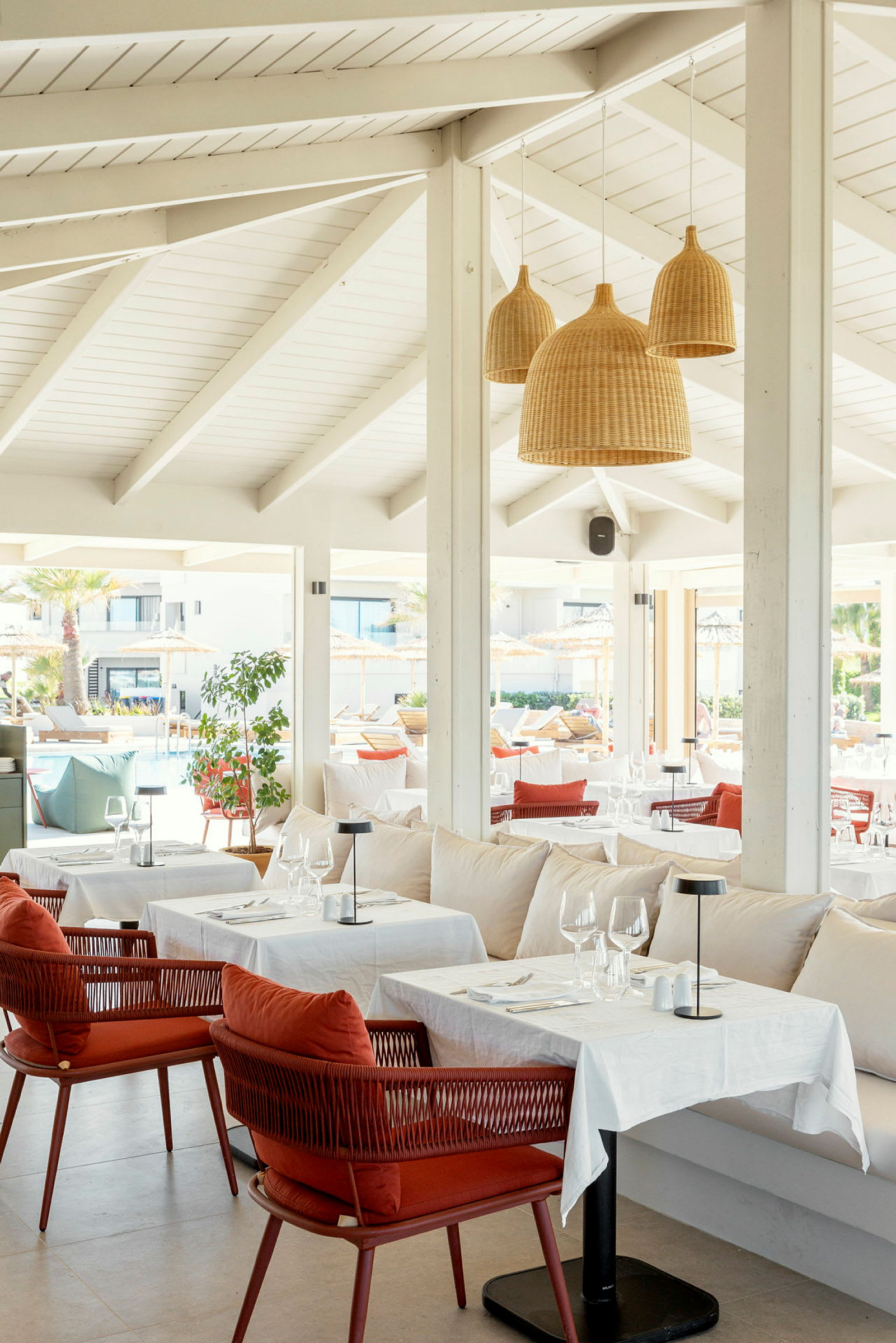 Interiör av en restaurang med vita takbjälkar, hängande rottinglampor samt dukade bord och stolar. (AI-genererad text)
