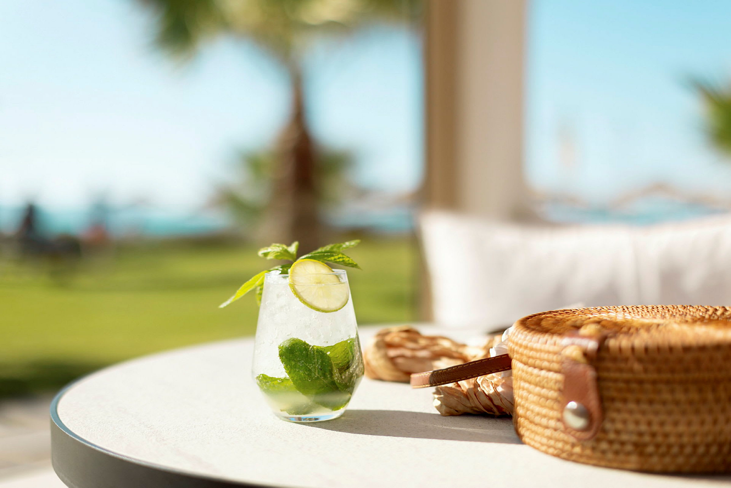 Glas med iskall drink innehållandes lime och mynta på ett bord, med en stråväska bredvid. (AI-genererad text)