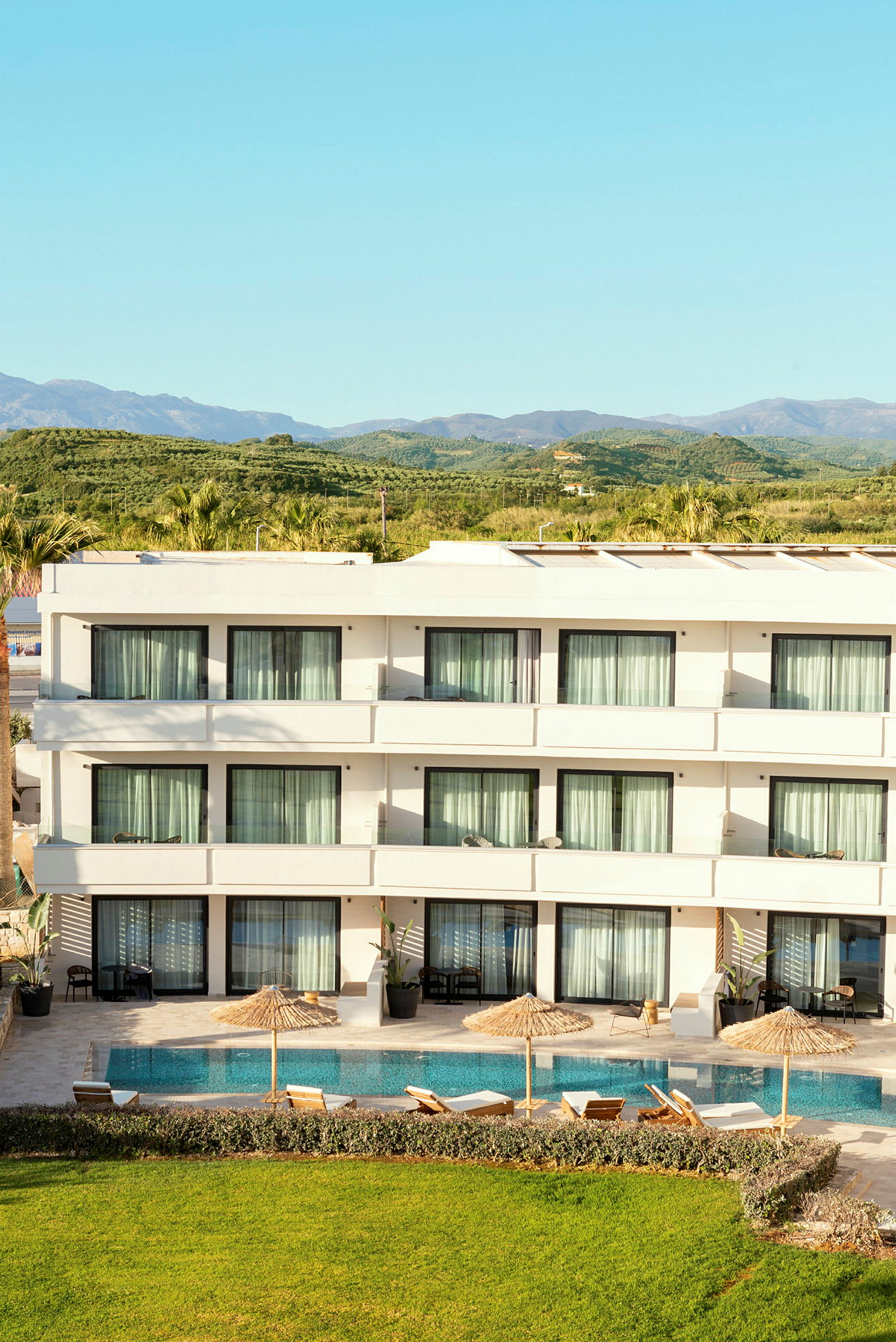 En hotellbyggnad med pool, solstolar och parasoller framför samt grönt landskap i bakgrunden. (AI-genererad text)