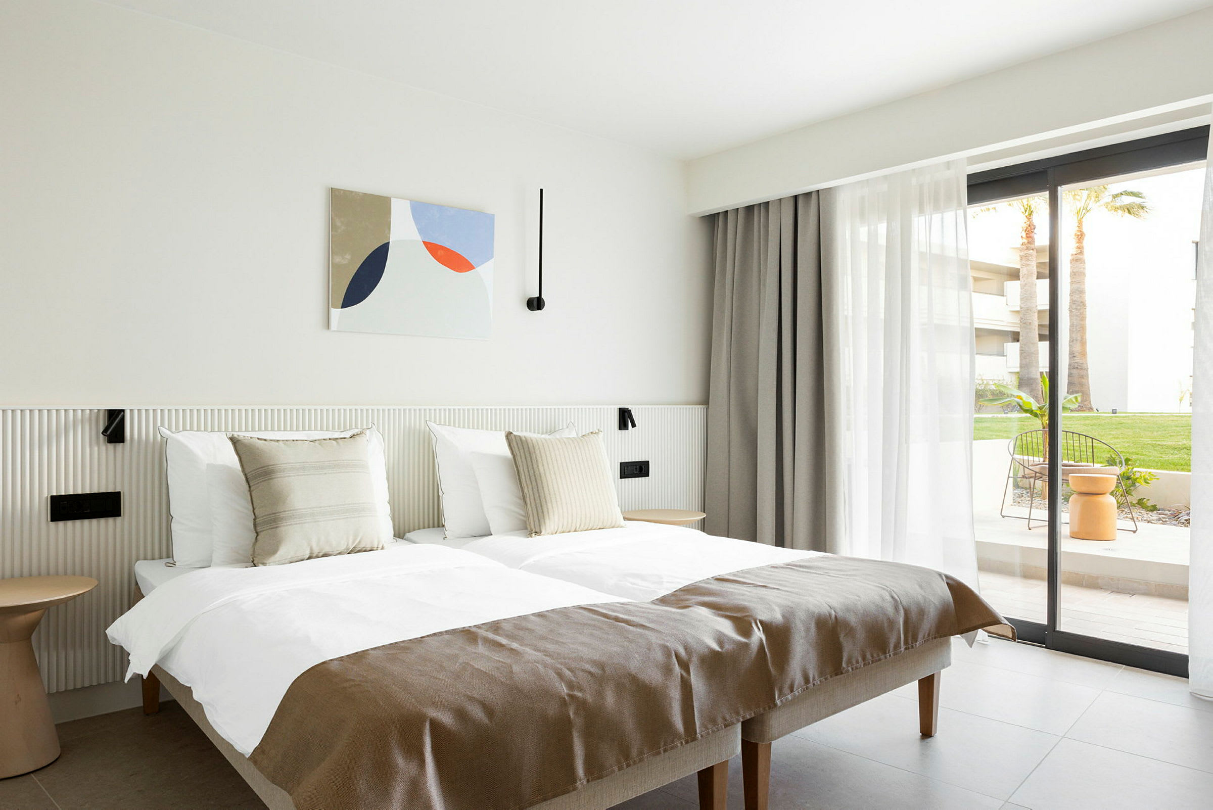 Ett ljust hotellrum med två enkelsängar, neutrala detaljer och en fönsterdörr mot en uteplats. (AI-genererad text)