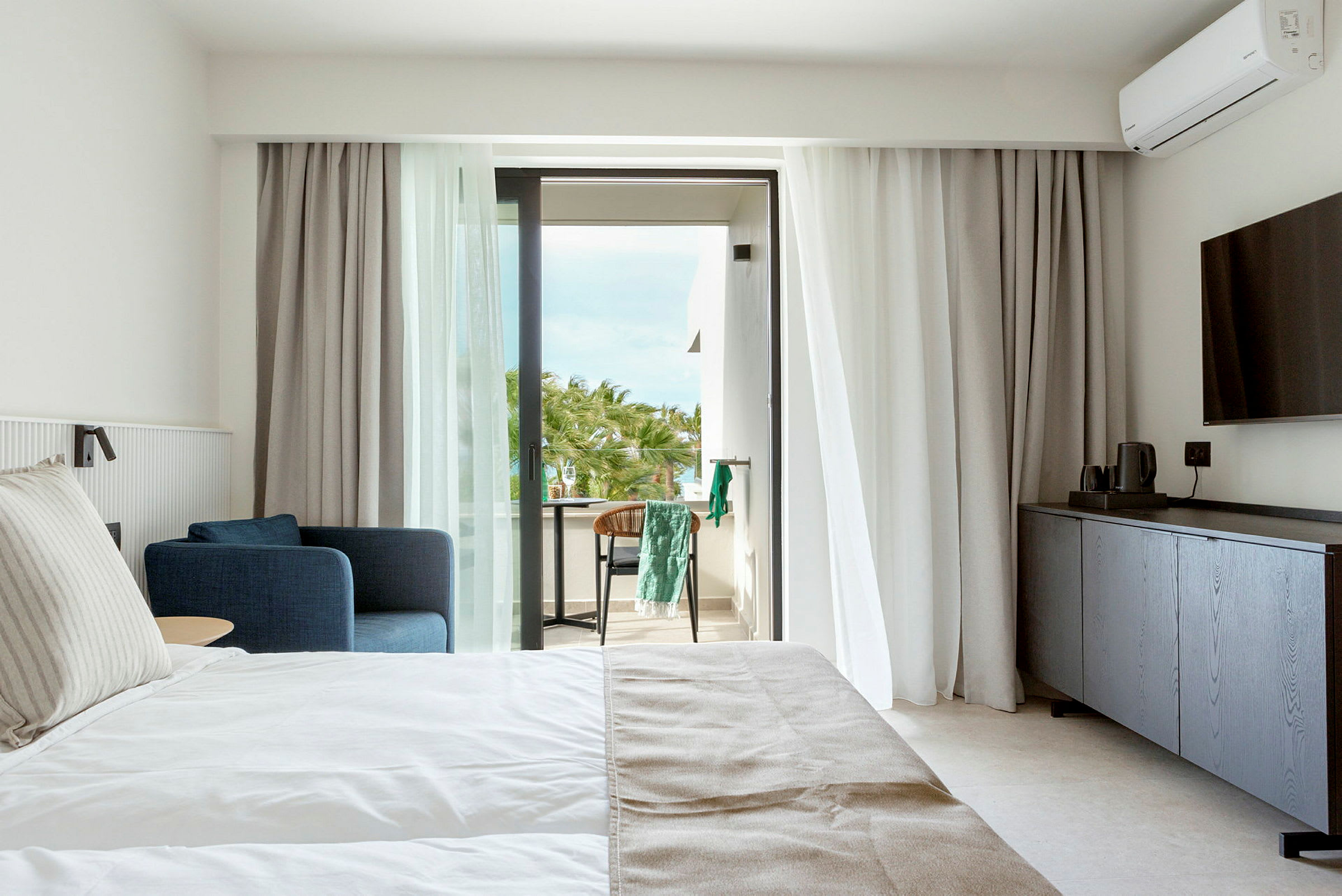 Hotellrum med säng, blå fåtölj och balkong med utsikt över palmer. (AI-genererad text)