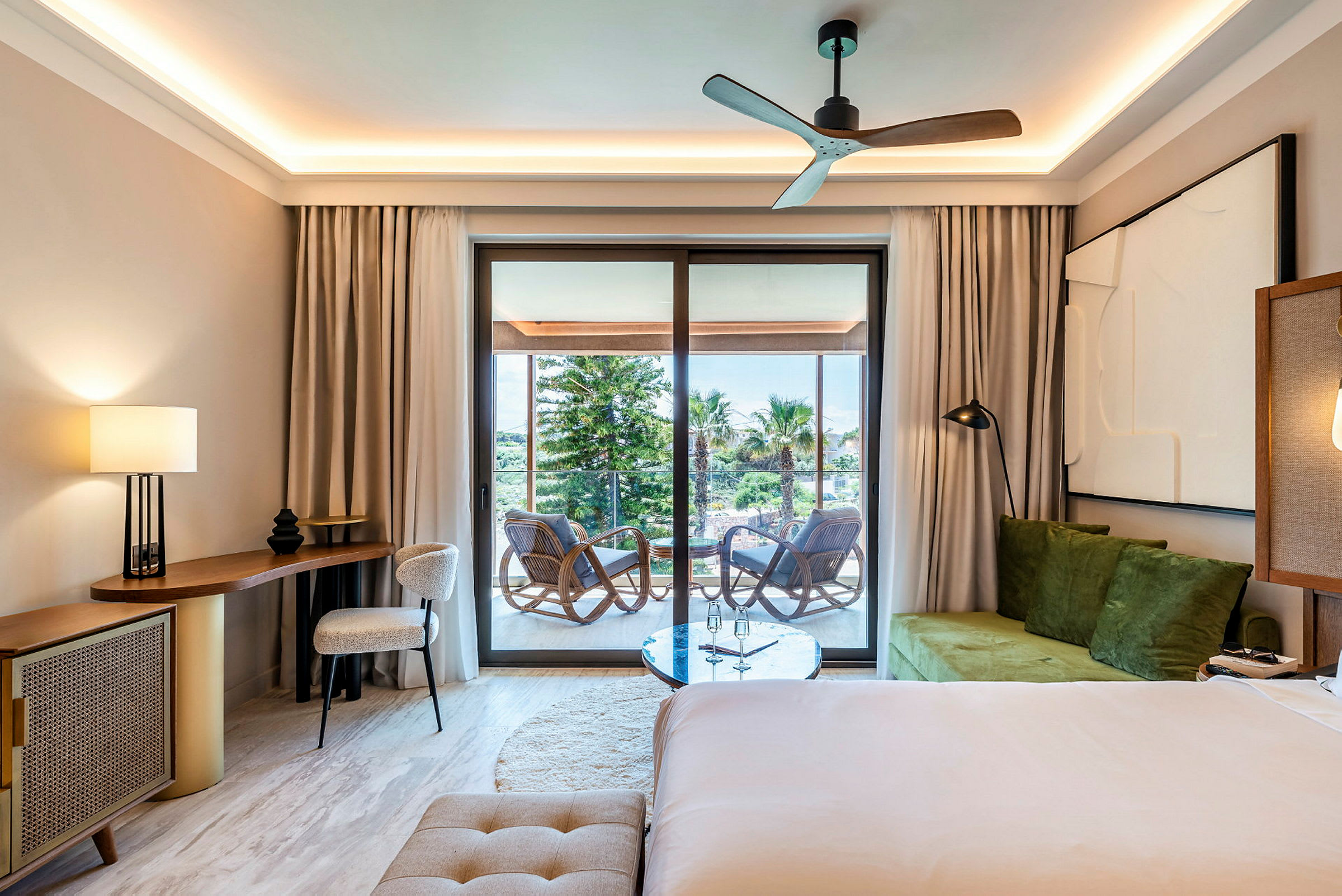 Ett hotellrum med modern inredning som inkluderar ett skrivbord, en stol, en säng och en balkong med möbler och utsikt över träd och himmel. (AI-genererad text)