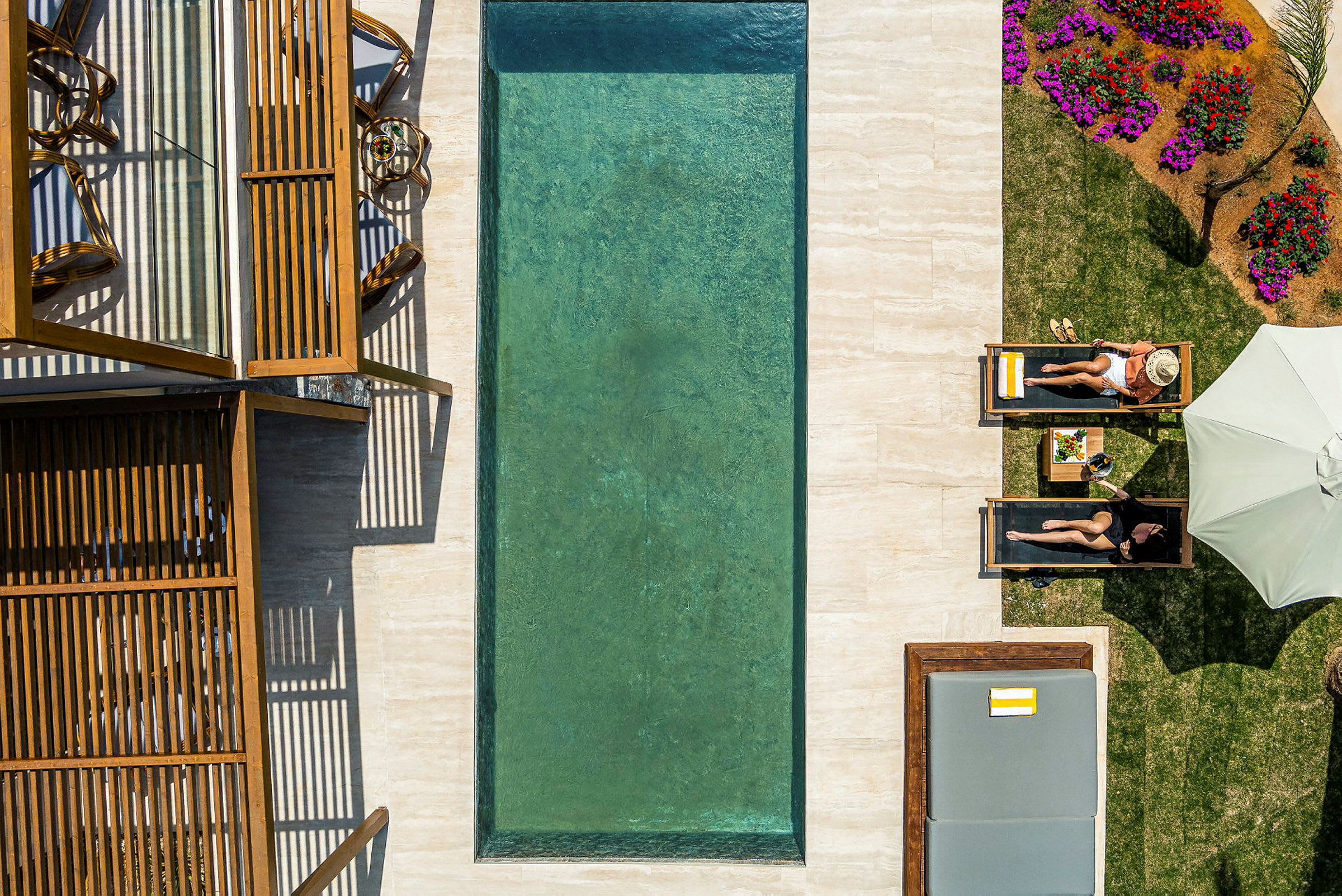 En pool med grönt vatten omgiven av solstolar, parasoller och blomsterbädd. Två personer ligger på solstolar vid sidan av poolen. (AI-genererad text)