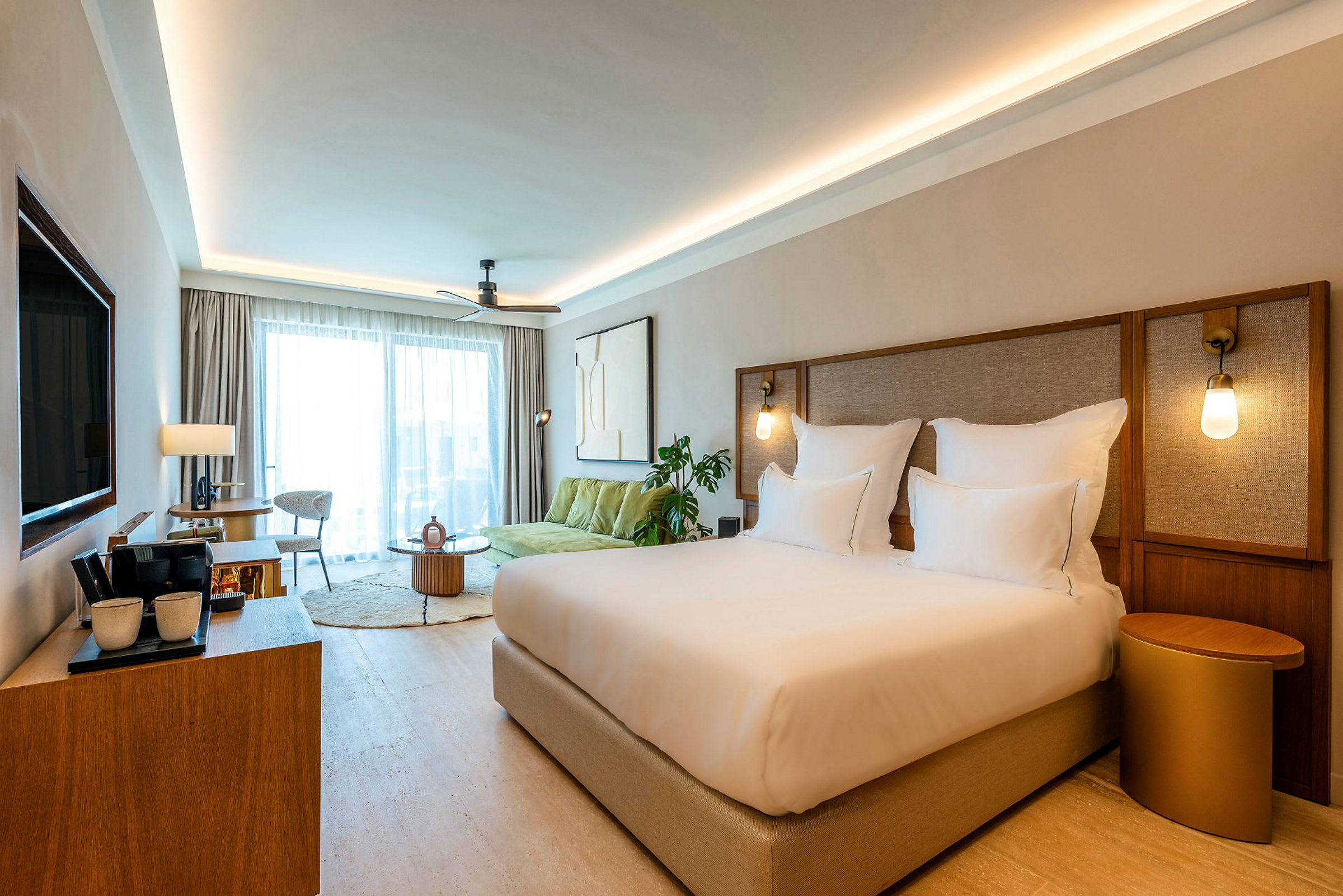 Ett elegant hotellrum med en stor säng, sittgrupp och stora fönster med gardiner. Vägglampor ger varm belysning. (AI-genererad text)