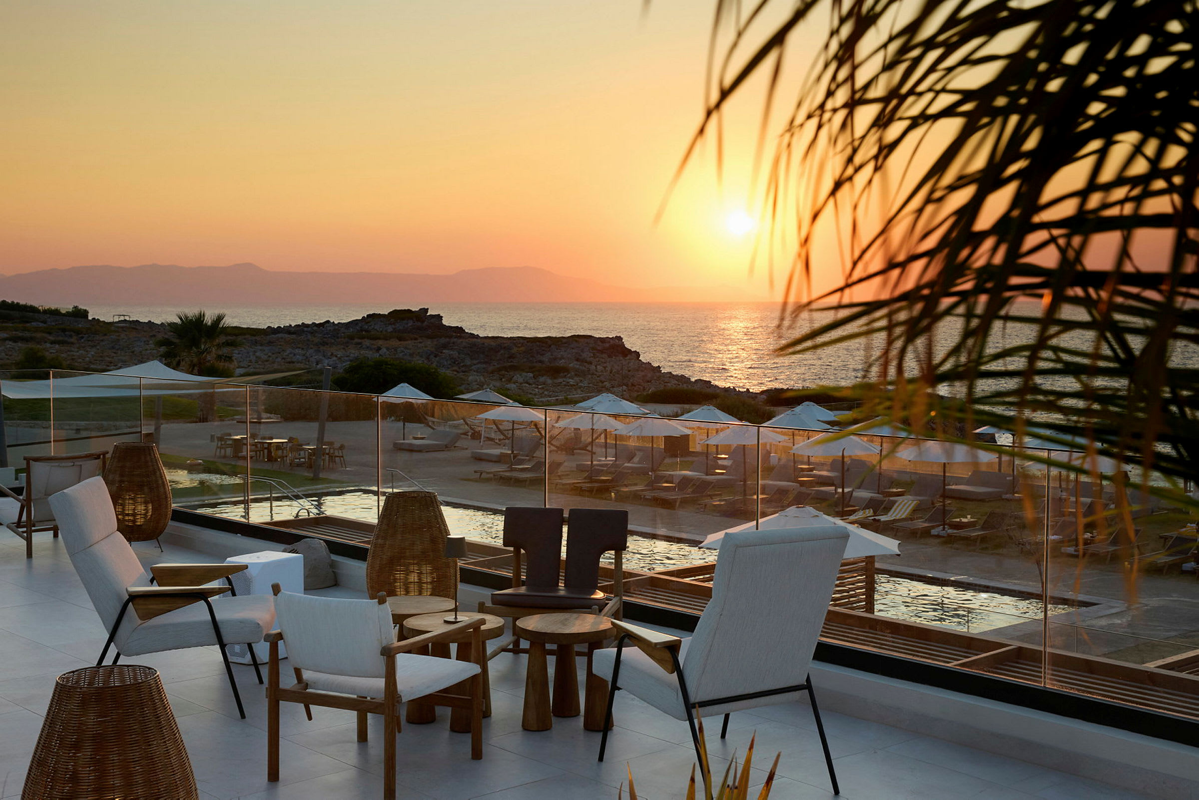 Terrass med sittplatser i förgrunden, med utsikt över en strand med solstolar och parasoller vid solnedgång. (AI-genererad text)