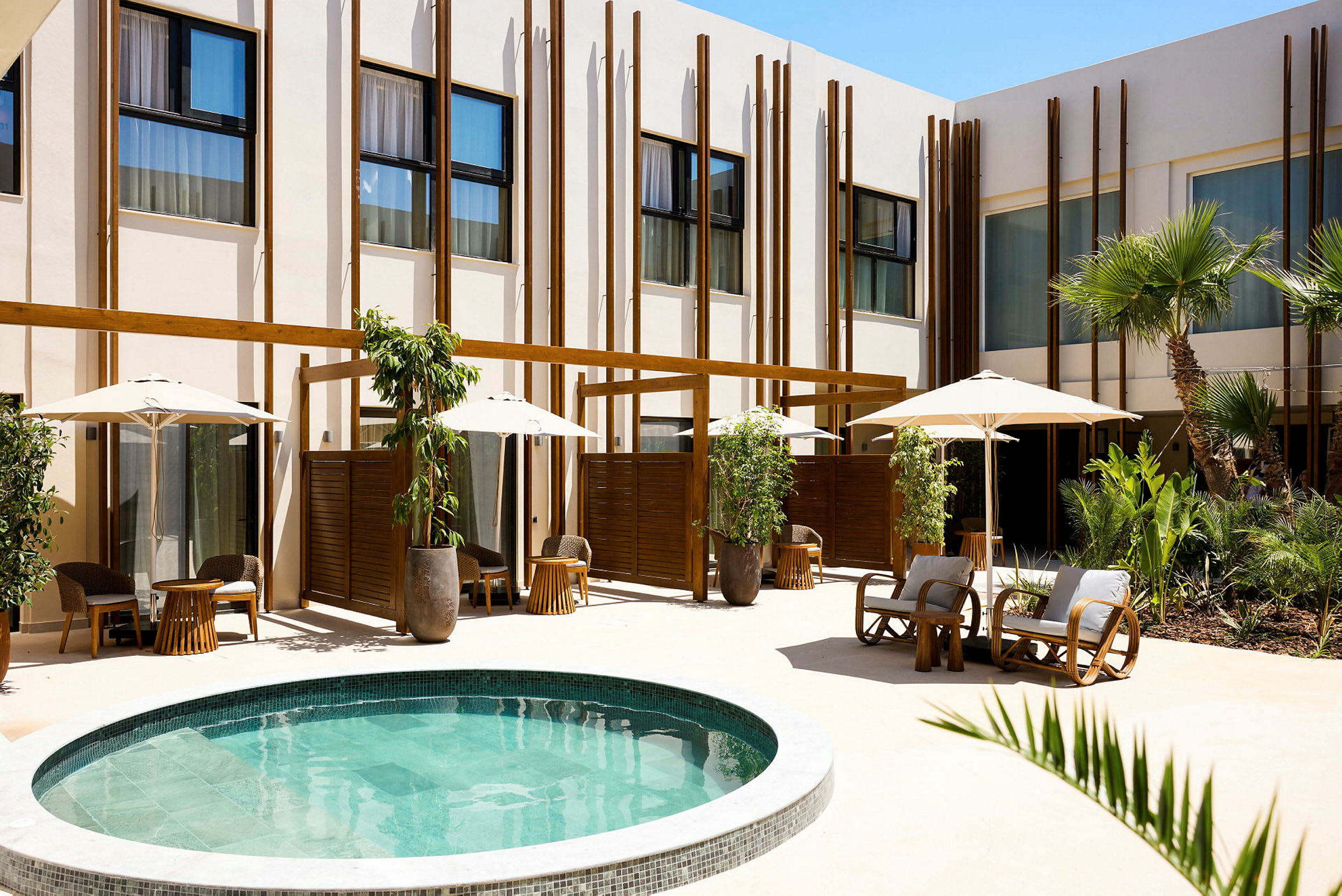 Ett modernt hotellområde med en liten rund pool omgiven av grönska, solstolar och parasoller. (AI-genererad text)