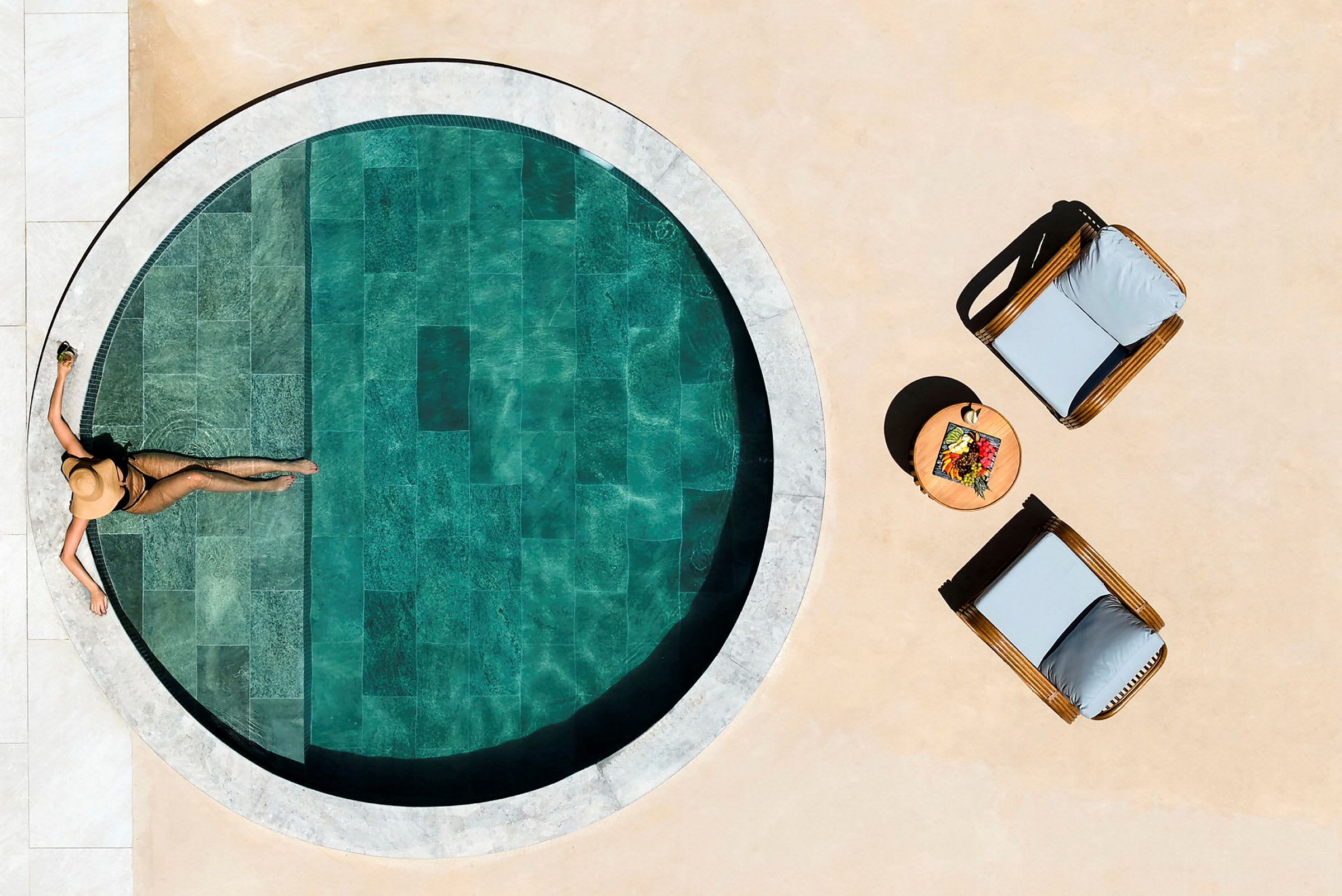 En person i solen vid en liten rund pool, med två solsängar och ett bord med frukt på sidan. (AI-genererad text)
