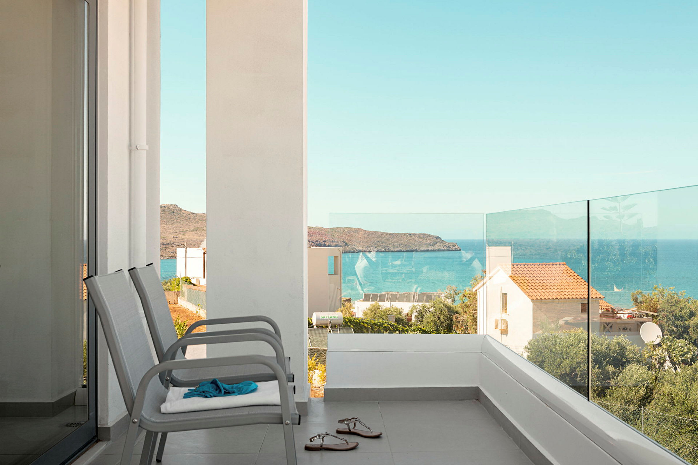 Utsikt från en balkong med glasräcke mot havet och omgivande hus. (AI-genererad text)