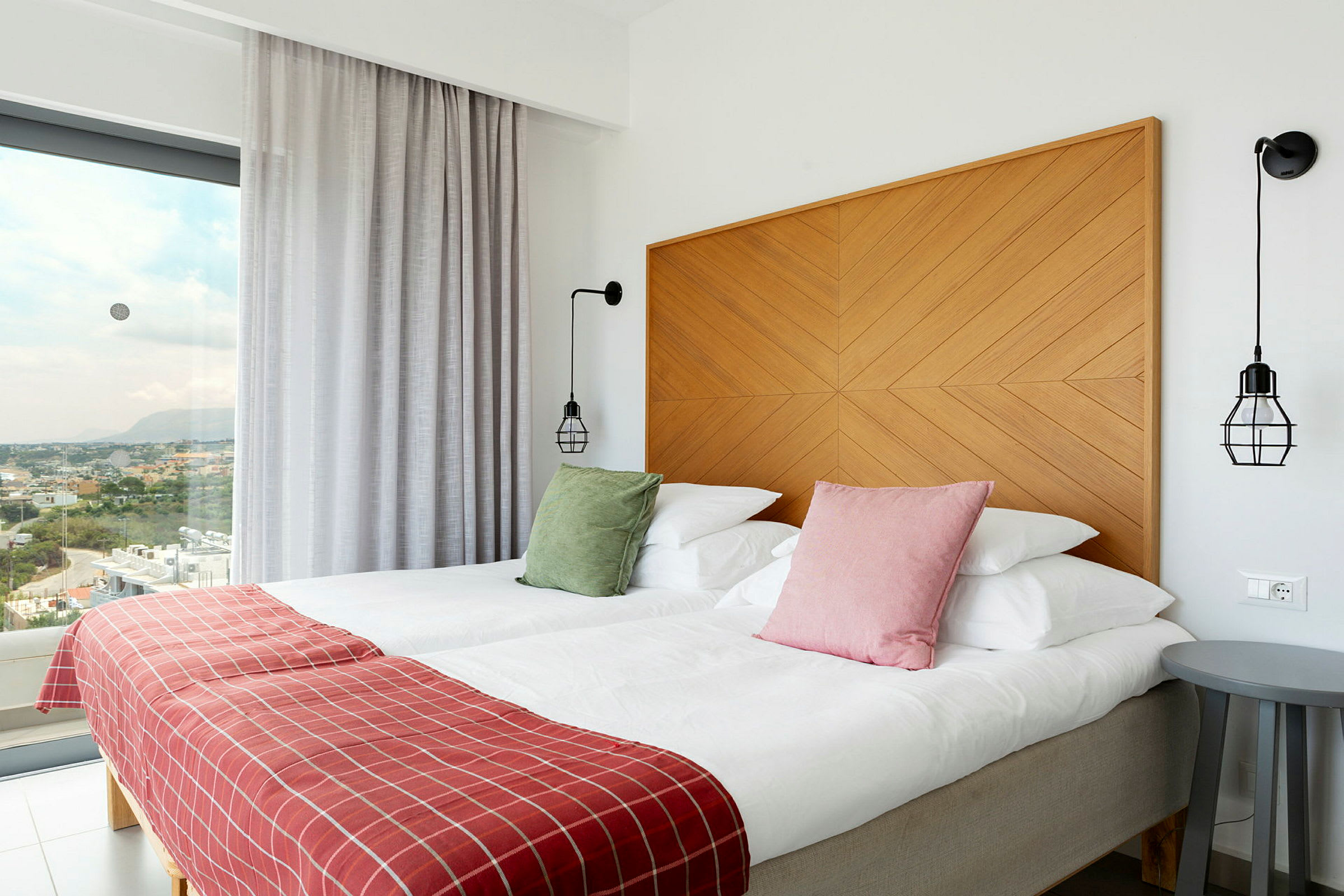 Hotellrum med två bäddar, färgglada kuddar, rödplädar och modernt inrett, med utsikt genom stora fönster. (AI-genererad text)