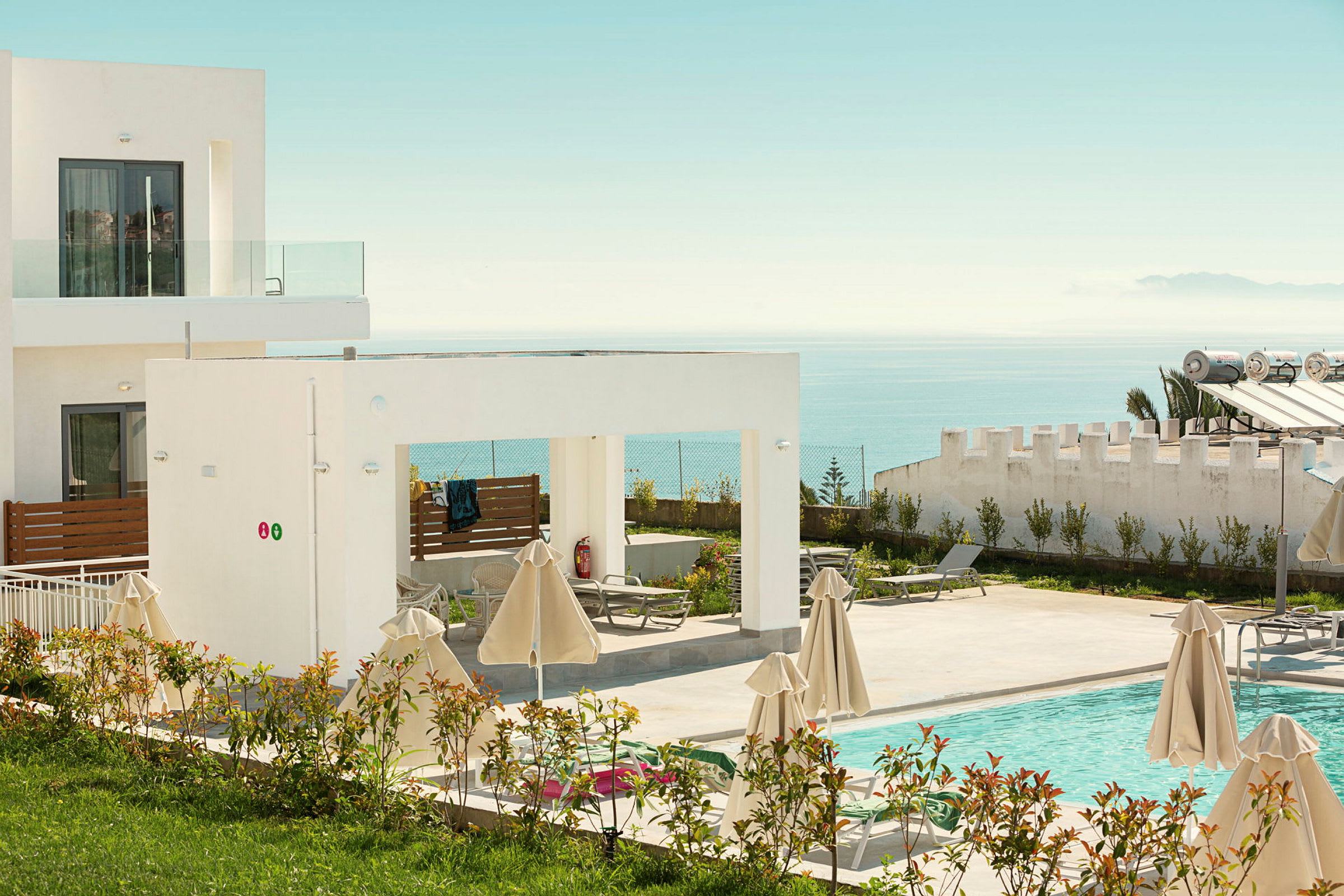 Bild av ett hotell med modern arkitektur, en pool och solstolar omgivna av parasoller samt utsikt över havet. (AI-genererad text)