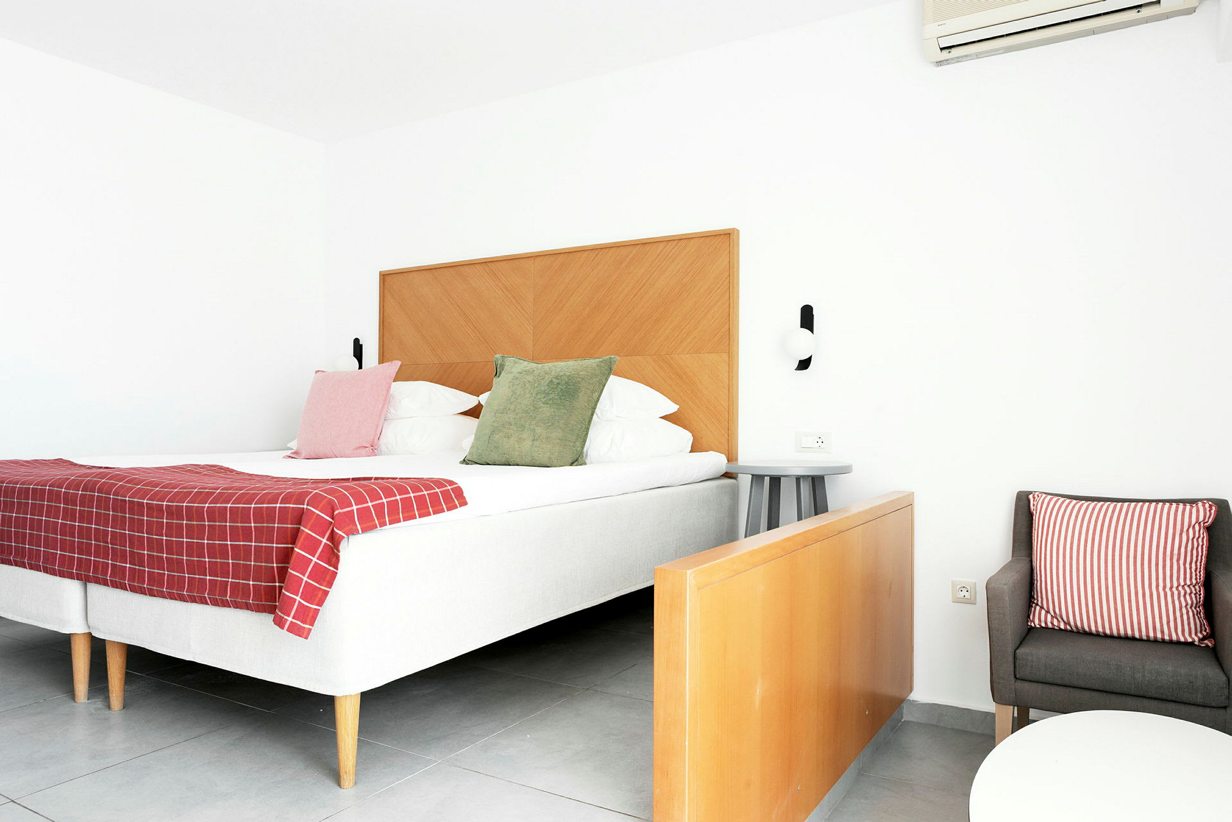Ett hotellrum med en dubbelsäng, träpanel på sänggaveln, rödrutigt täcke, färgade kuddar och en sittgrupp med stol och bord. (AI-genererad text)