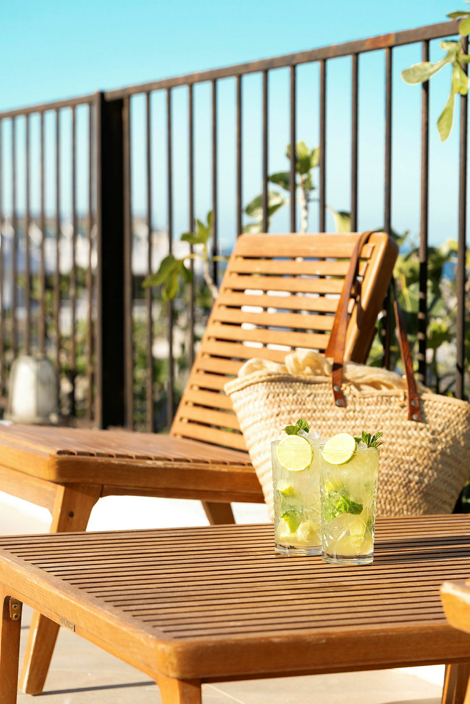 Två glas med lime- och myntadryck på ett träbord framför en solstol och en strandväska. (AI-genererad text)