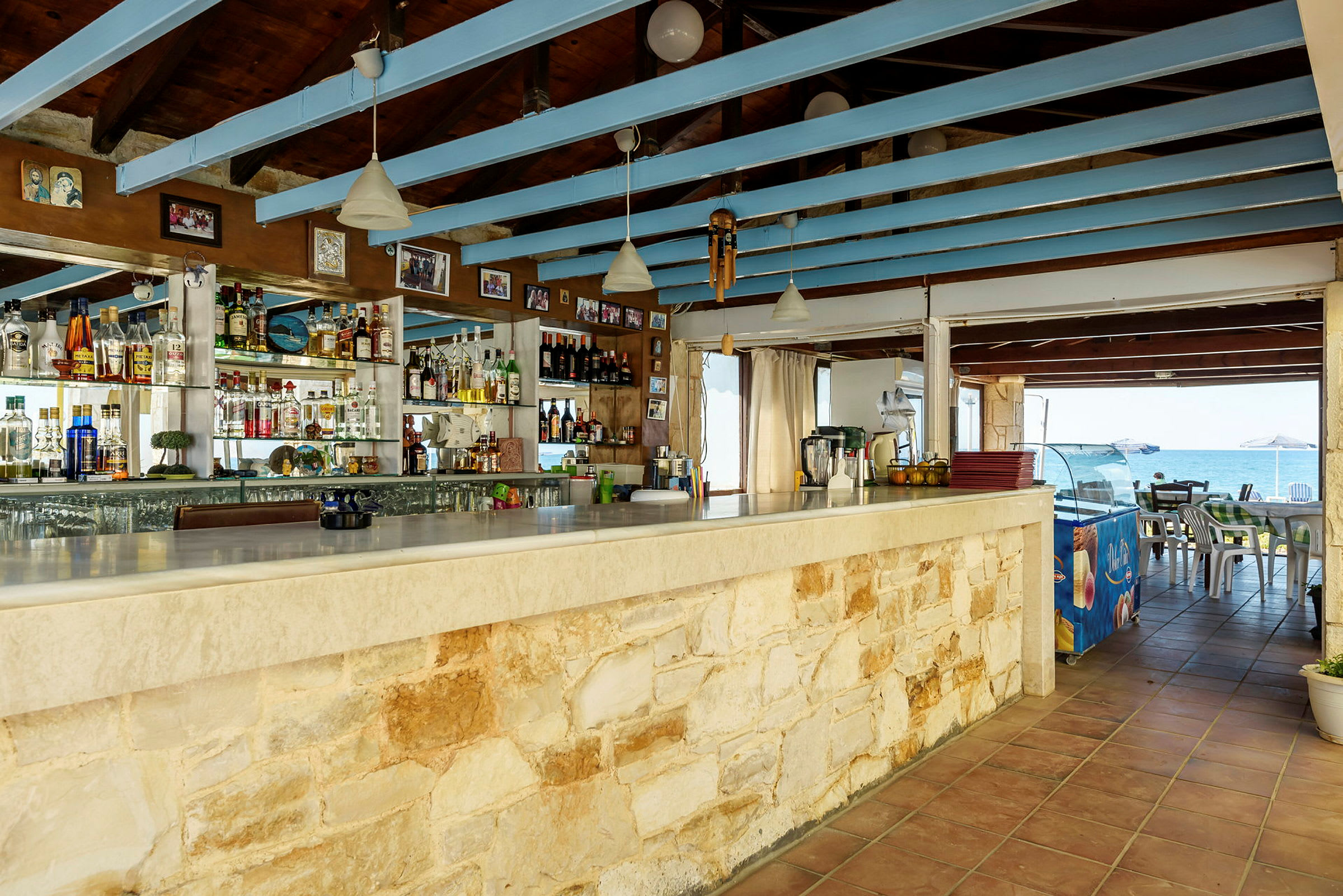 En taktäckt strandbar med en stenklädd bardisk och hyllor fulla av flaskor och glas, med havsutsikt i bakgrunden. (AI-genererad text)
