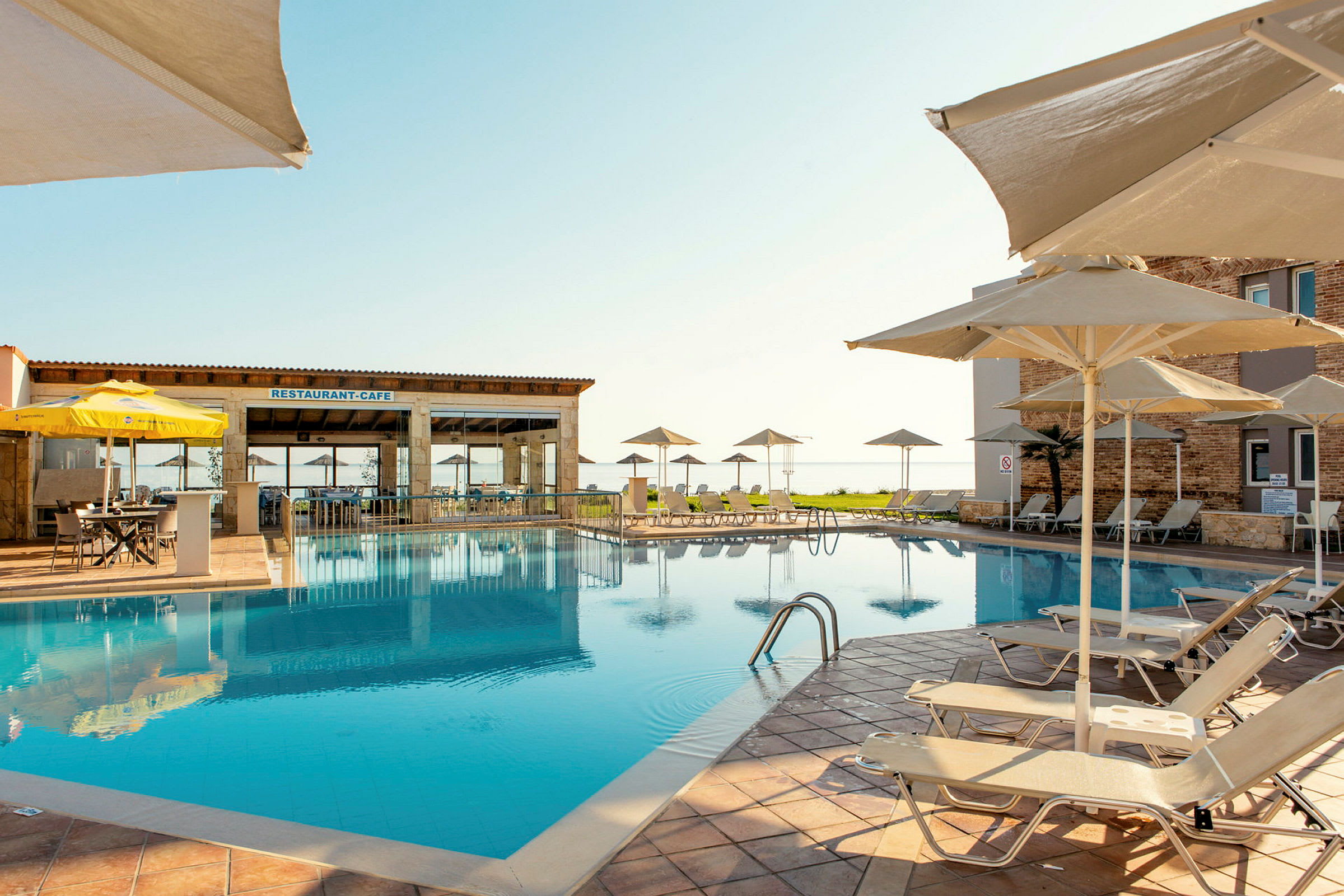 En utomhuspool med solstolar och parasoller vid ett hotell. En restaurang byggnad syns i bakgrunden. (AI-genererad text)