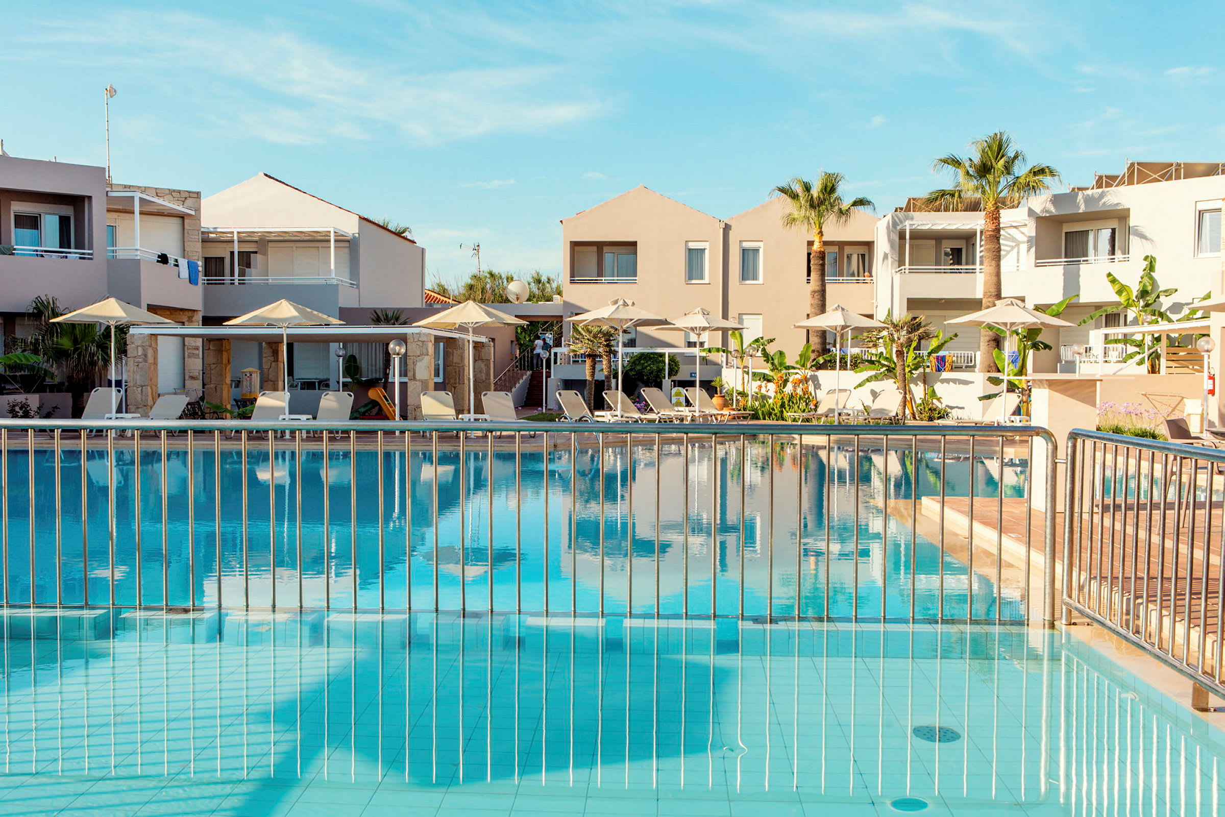 En pool med solstolar och parasoller runt, framför semesterlägenheter. (AI-genererad text)