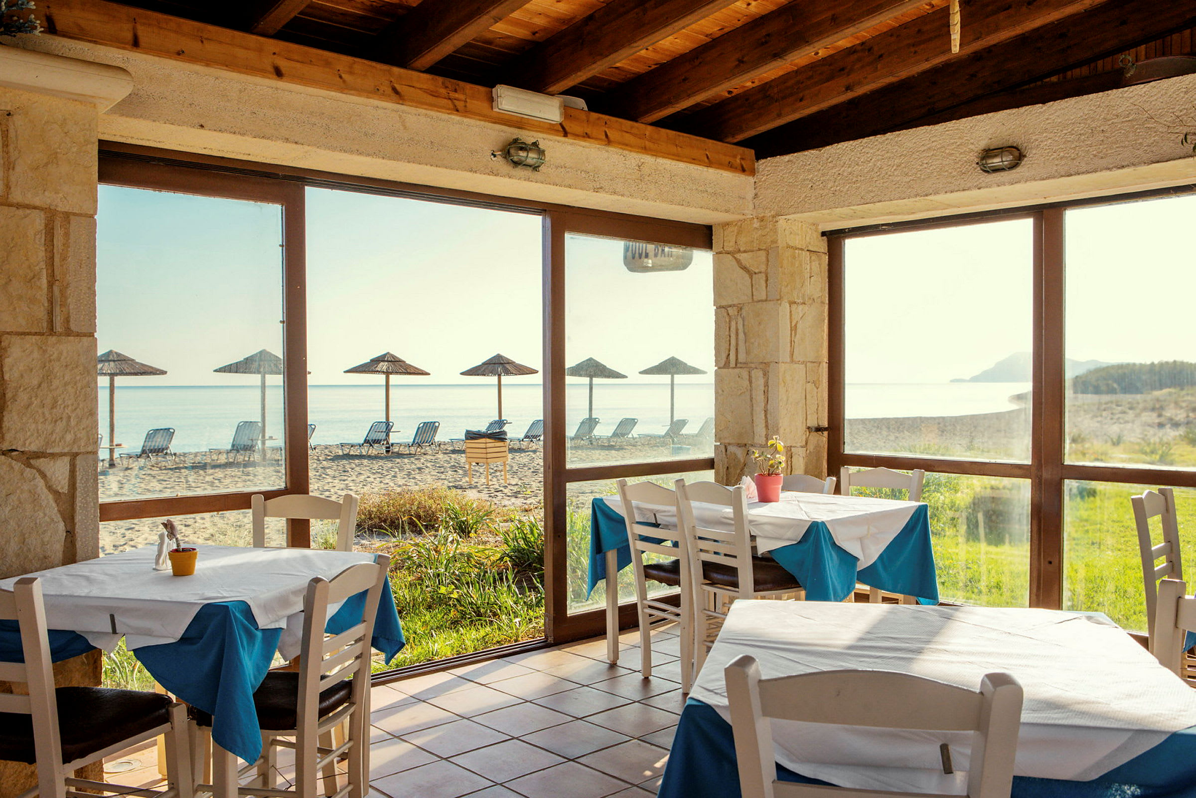 Restaurang med utsikt över stranden, med bord och stolar inomhus samt parasoller och solstolar utomhus. (AI-genererad text)