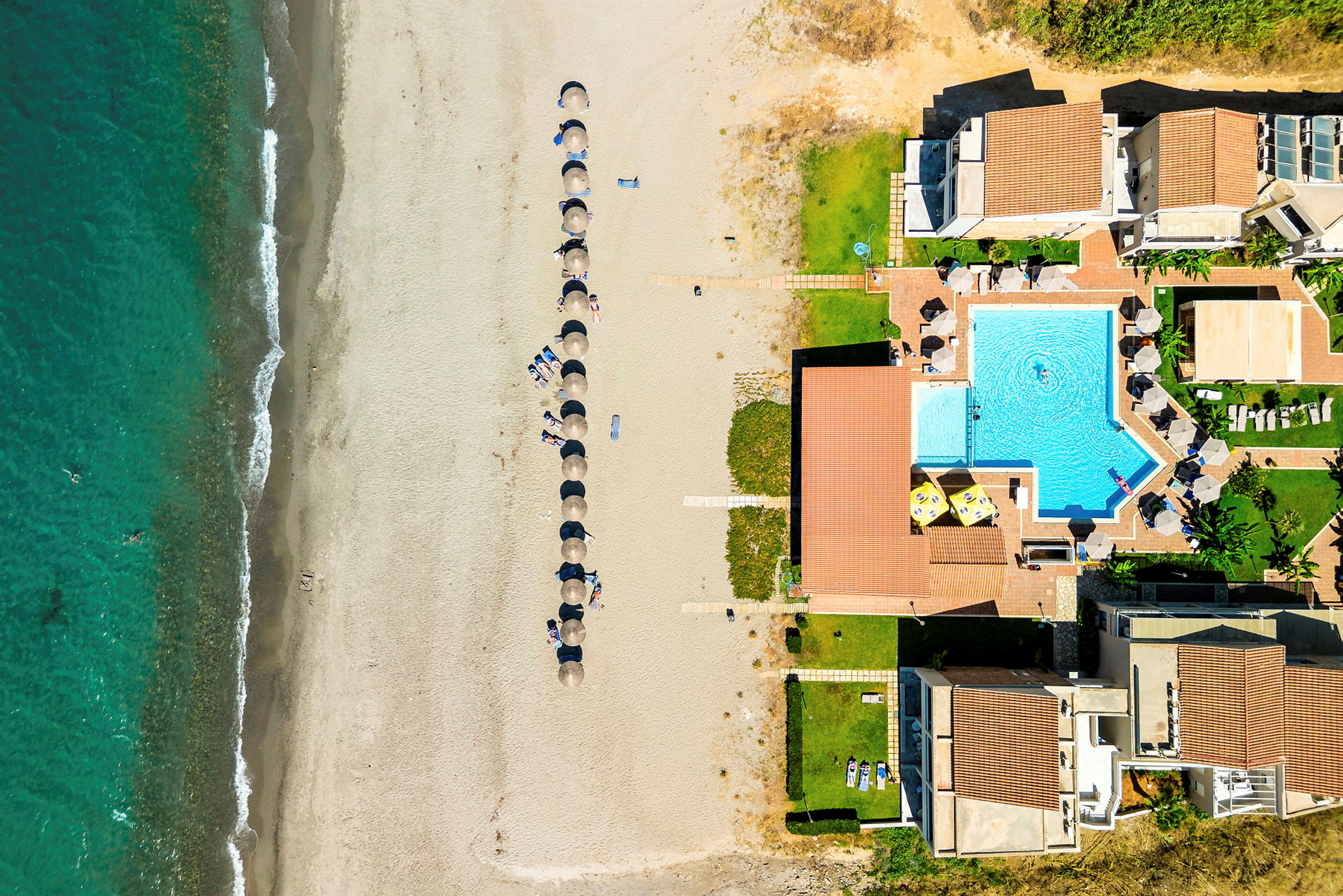 Flygfoto över en strand med parasoller och en pool nära byggnader. (AI-genererad text)