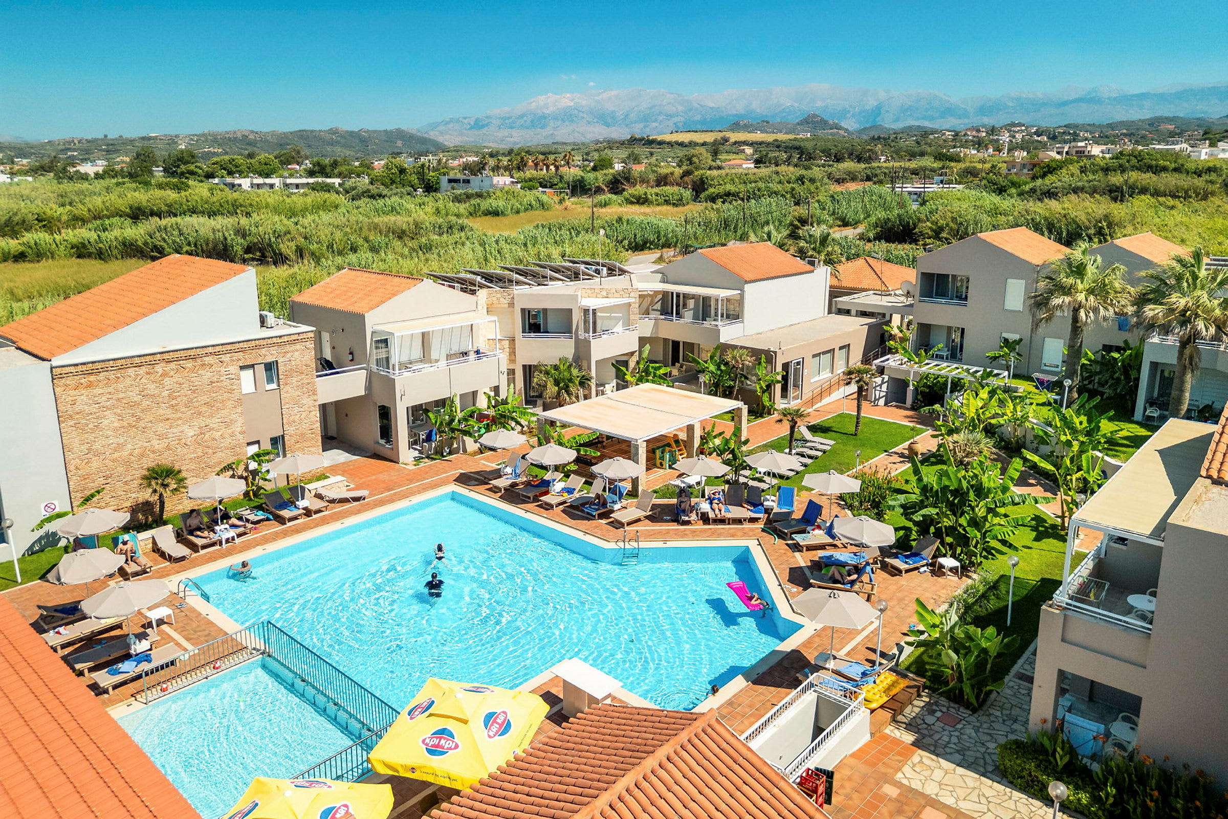 Hotellområde med en pool omgiven av solstolar och parasoller, med grönska och byggnader i bakgrunden. (AI-genererad text)