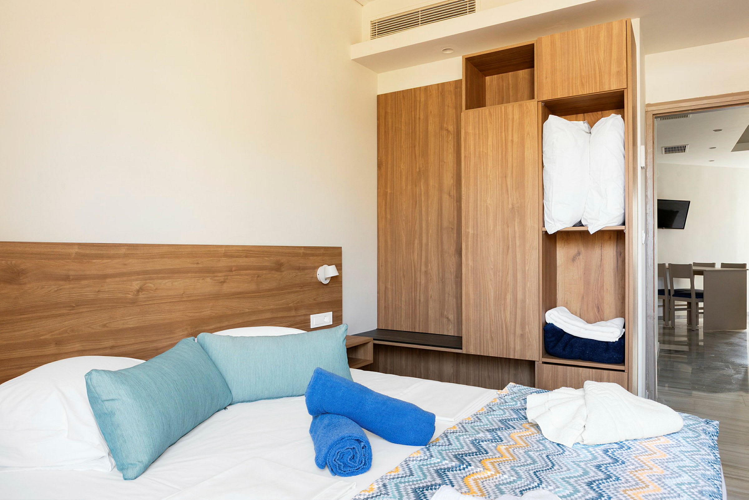 Ett hotellrum med en säng som har blå kuddar, blå och vita handdukar samt ett färgglatt mönstrat överkast. I bakgrunden syns en träfärgad garderob och en dörr mot ett annat rum. (AI-genererad text)