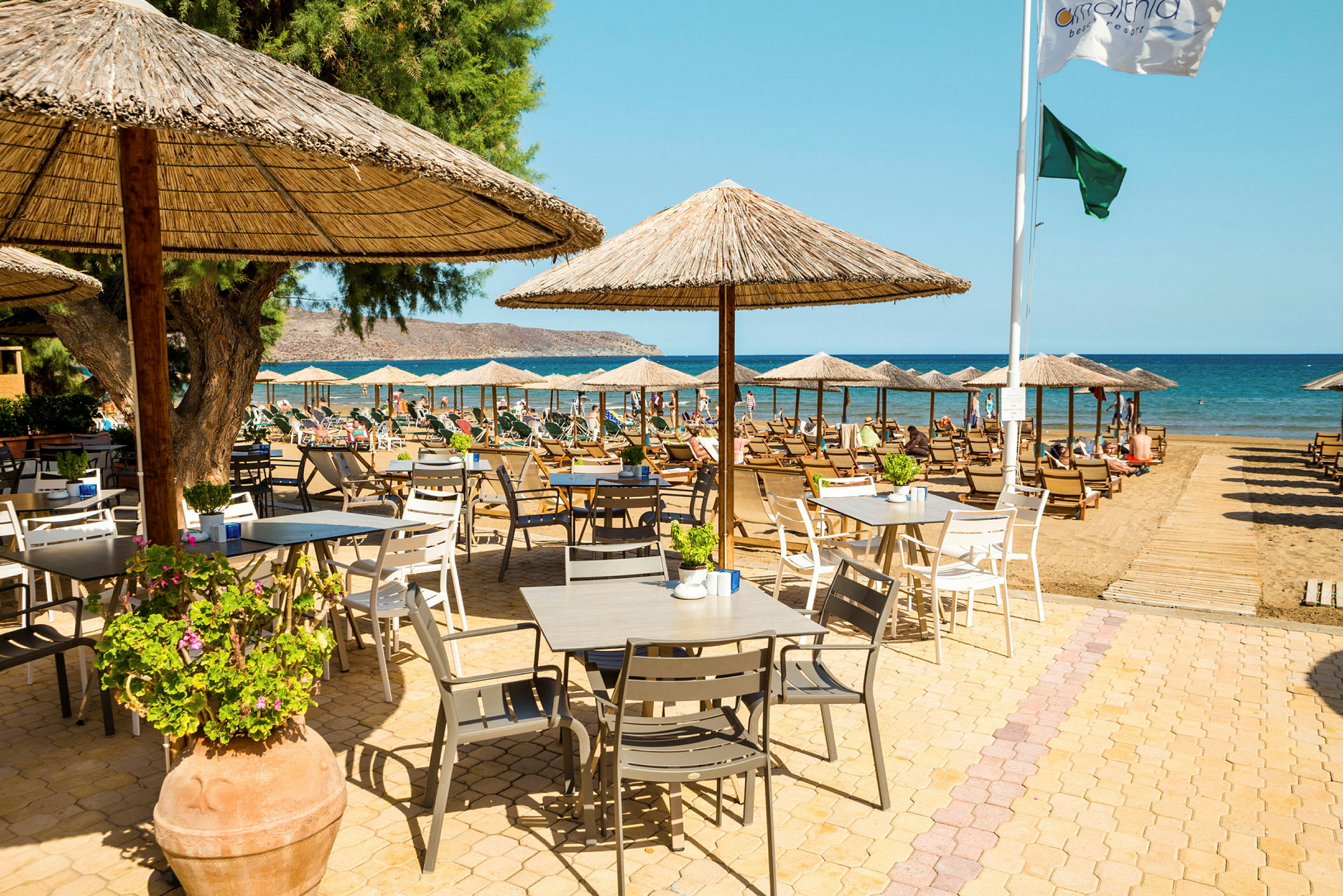 Bild av ett område vid stranden med bord och stolar under parasoll samt solstolar nära havet. (AI-genererad text)