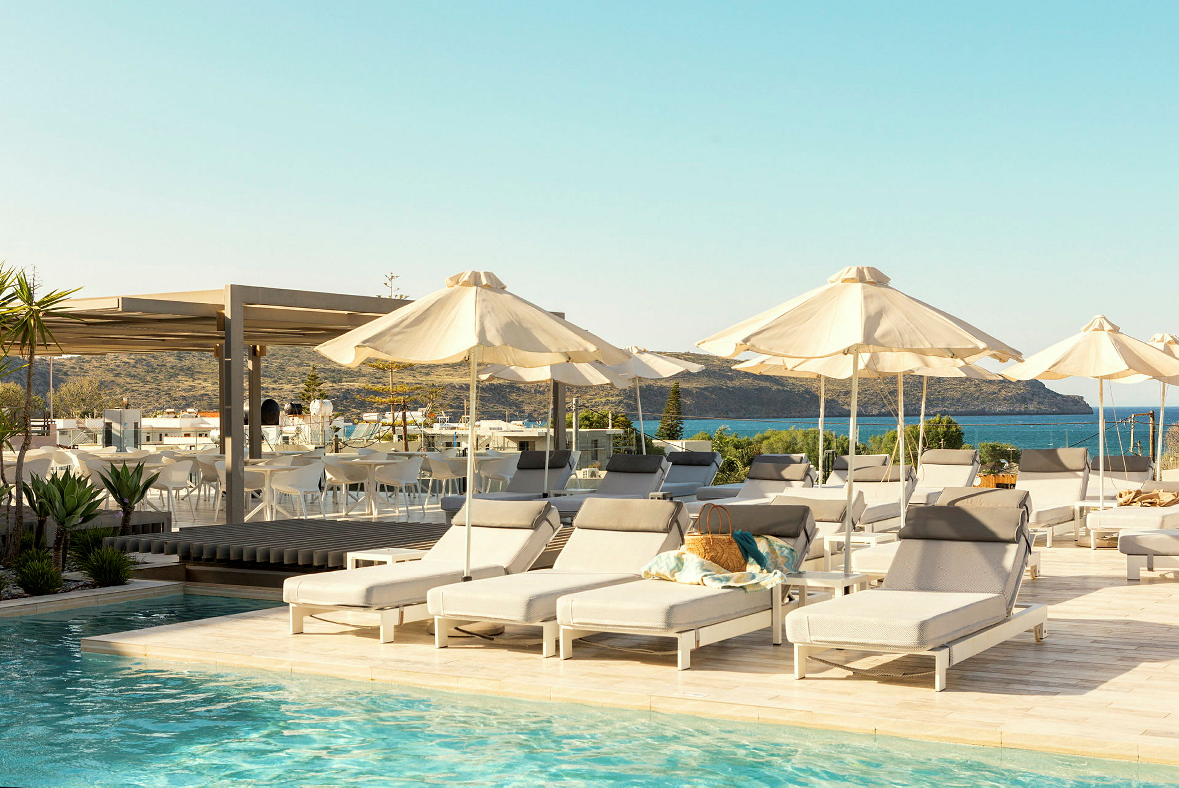 Bild av en pool med solstolar och parasoller runt omkring. I bakgrunden syns havet och en terrass med sittplatser. (AI-genererad text)
