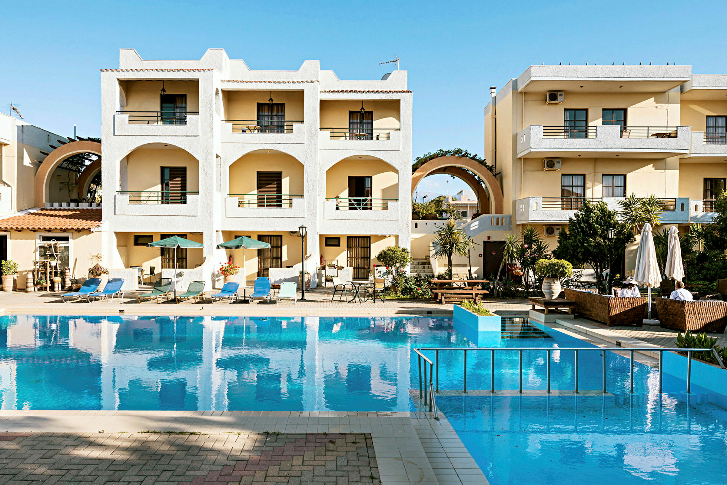 Ett hotellkomplex med balkonger och en pool framför byggnaden. Det finns solsängar och parasoller runt poolen. (AI-genererad text)